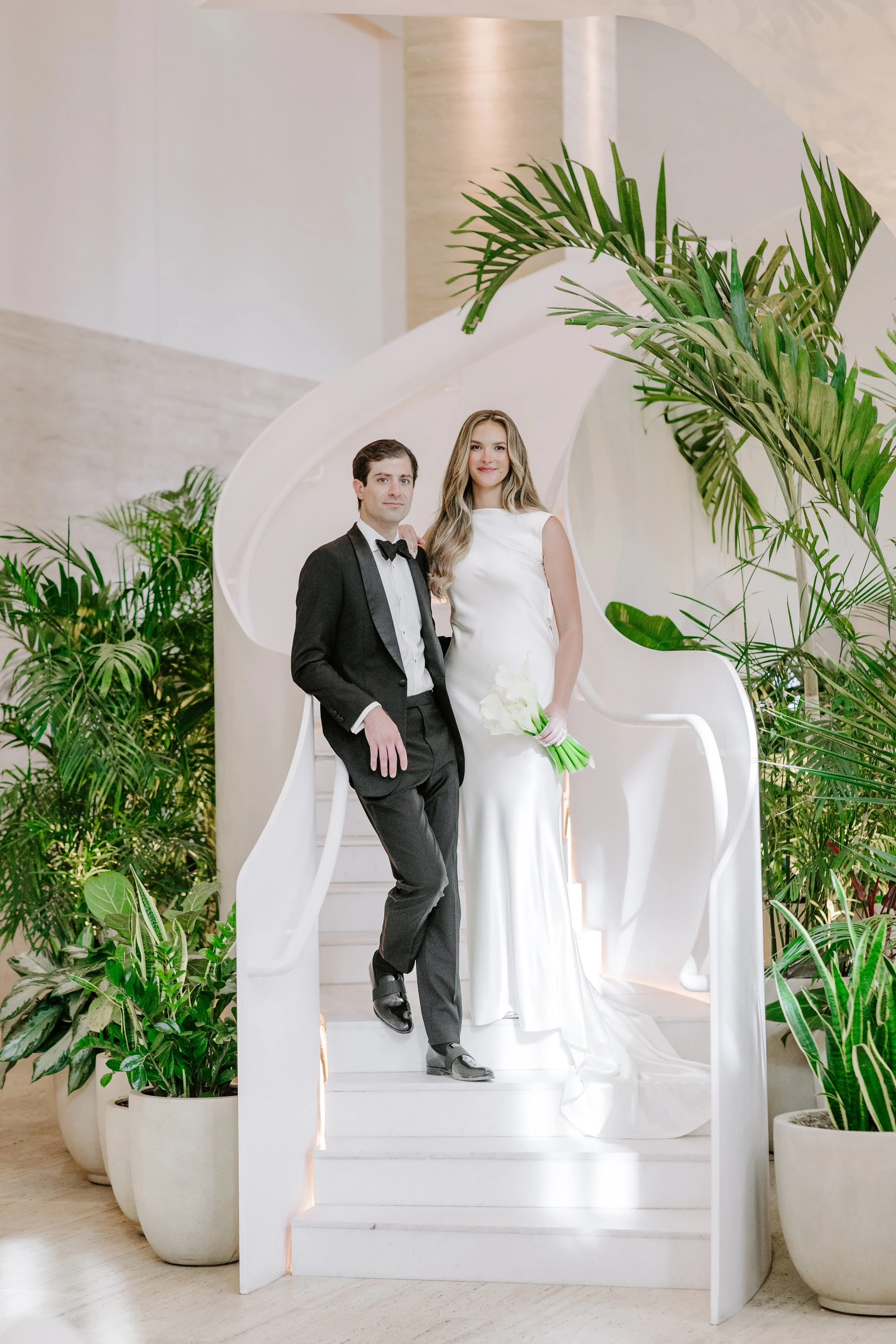 A couple dressed in formal wedding attire stands on a white staircase surrounded by large green potted plants. The man wears a tuxedo with a bow tie, and the woman wears a long white dress holding a bouquet of white flowers. The setting is bright and