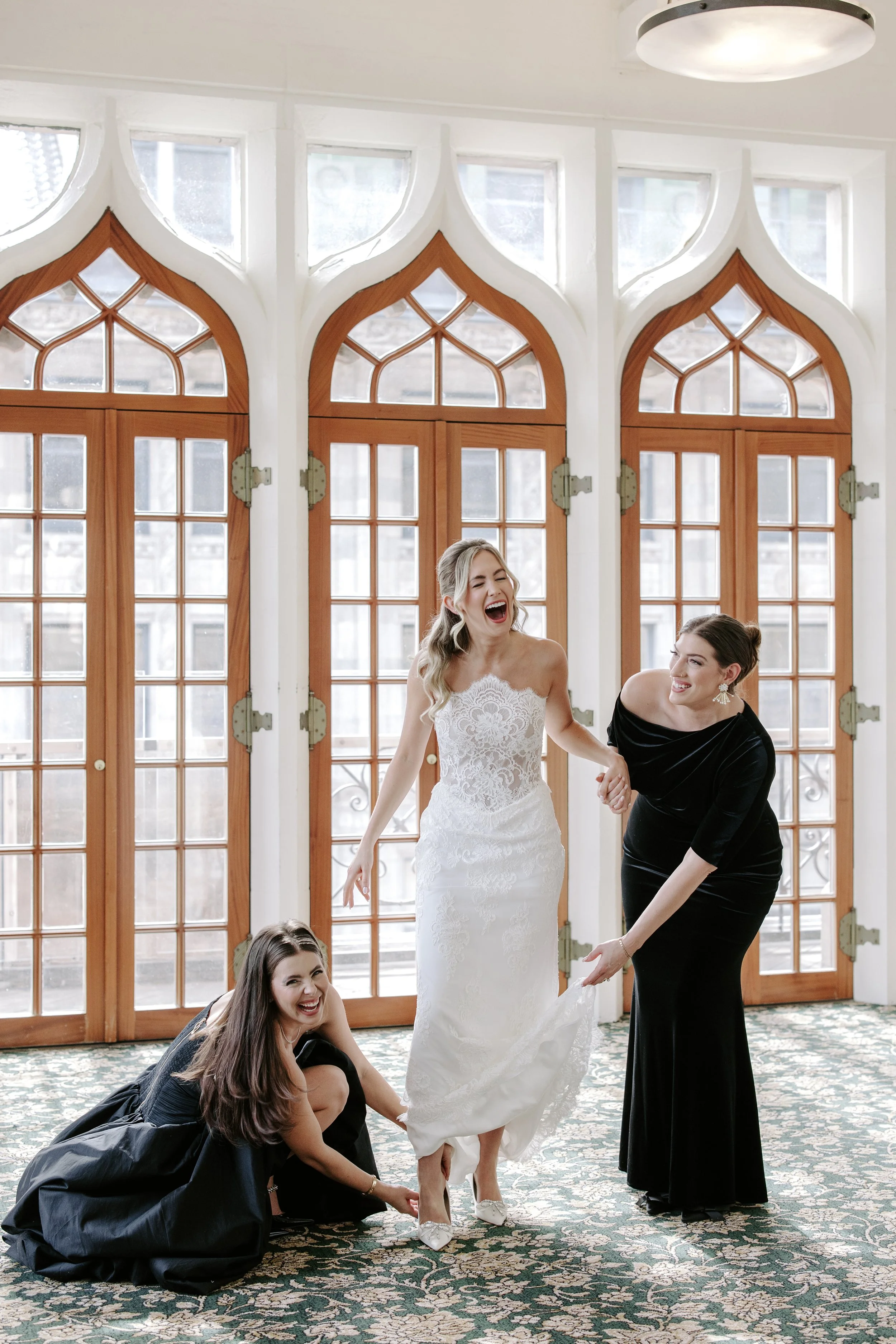 Four women, one bride in a white wedding dress, and three in black dresses, are laughing and smiling indoors with large wooden and glass doors in the background. Two women are crouched, helping the bride with her dress, while another woman holds the 