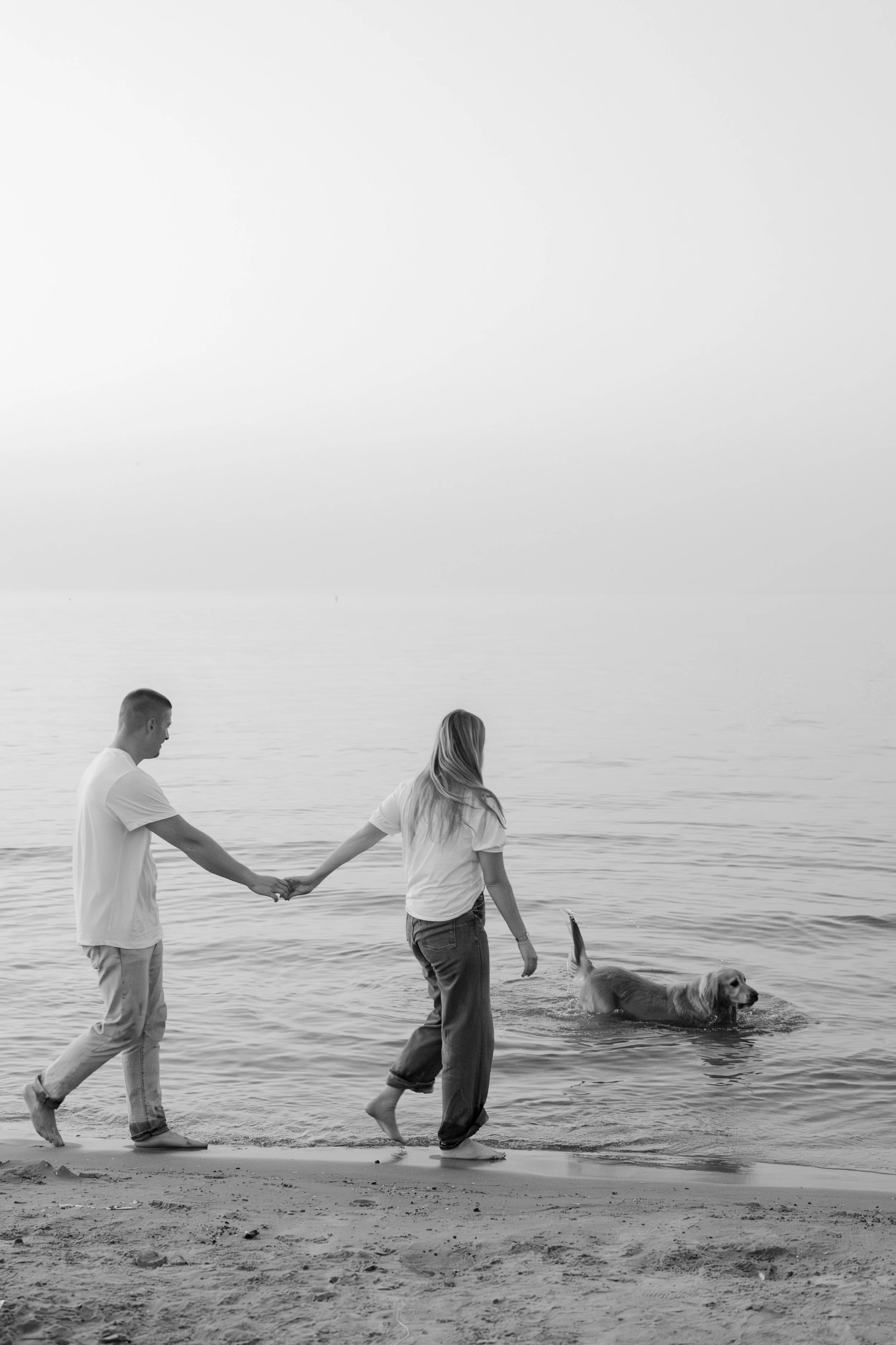 A couple holding hands and walking along the beach with their dog in the water, during daytime, in black and white.