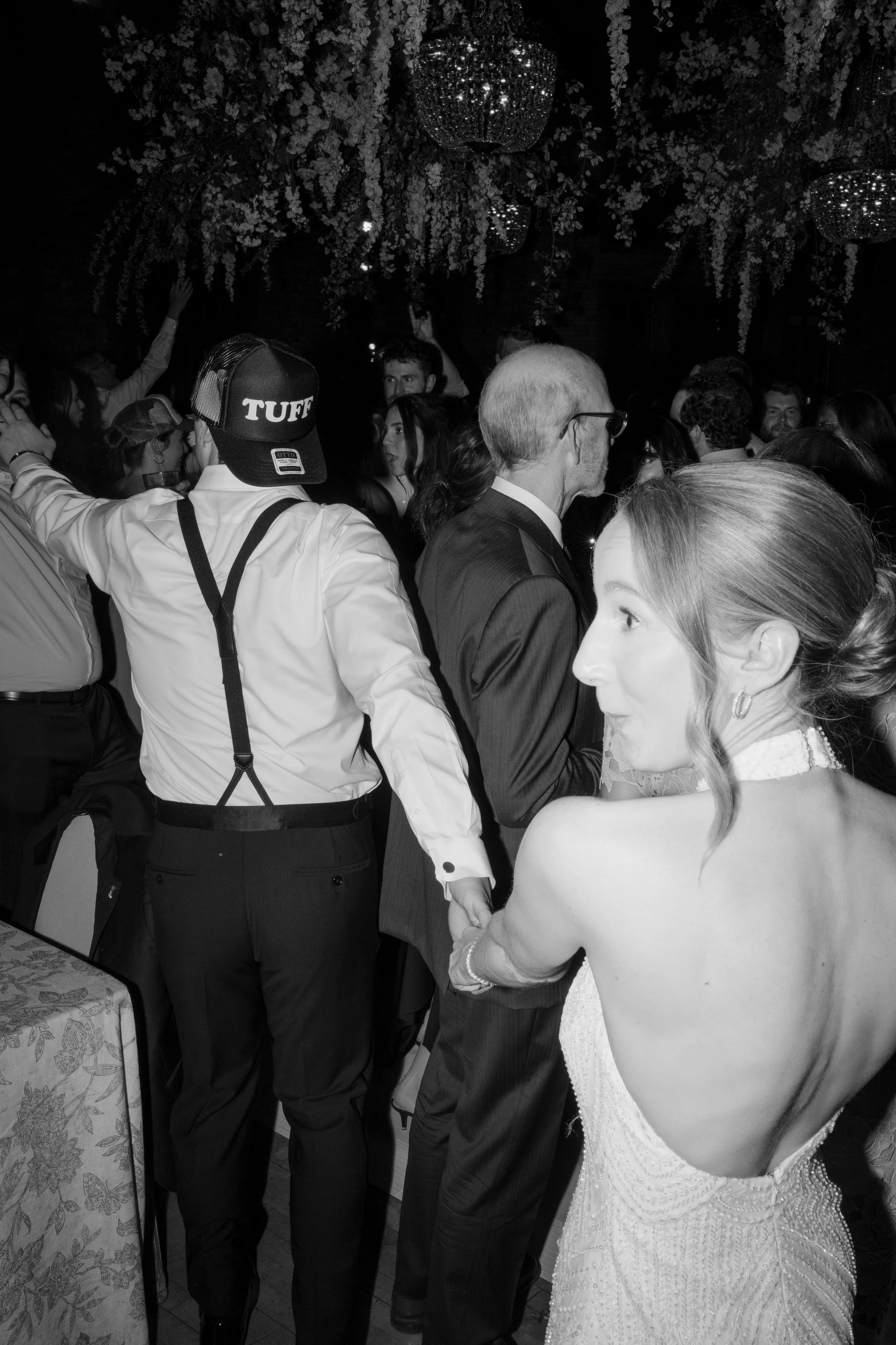 People dancing at a celebration event, with floral decorations hanging from the ceiling.