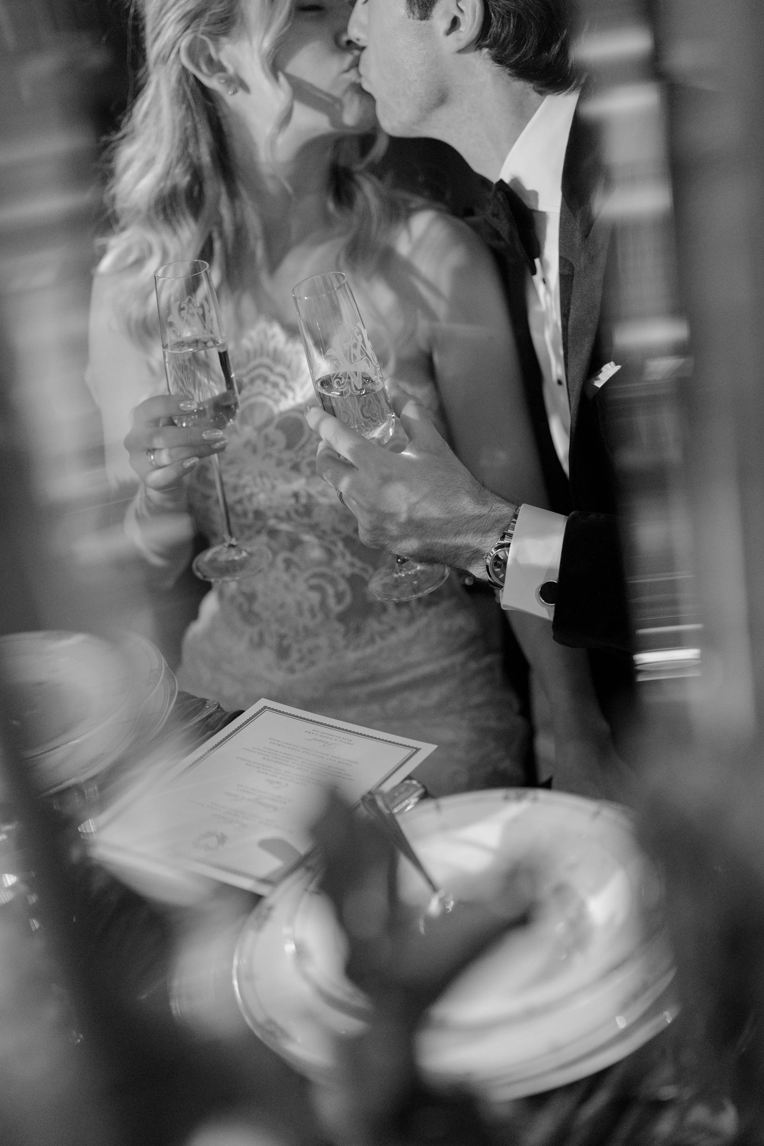 A black and white photo of a couple kissing, dressed in formal wedding attire, holding champagne glasses, with a menu or program on the table in front of them.
