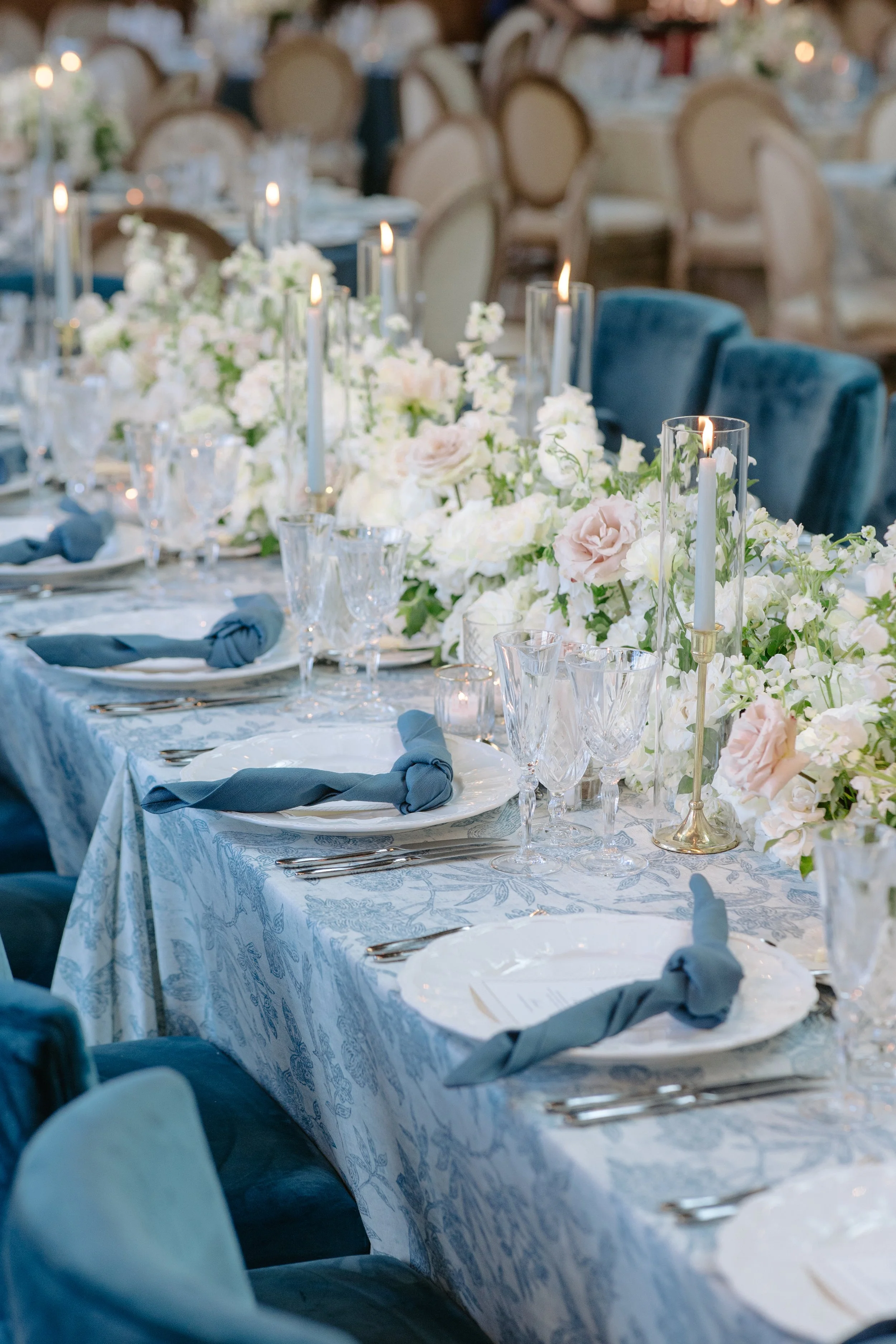 Elegant banquet table decorated with white and blush flowers, tall white candles in glass holders, blue napkins on white plates, and crystal glassware. Velvet chairs surround the table, with additional tables and chairs blurred in the background.
