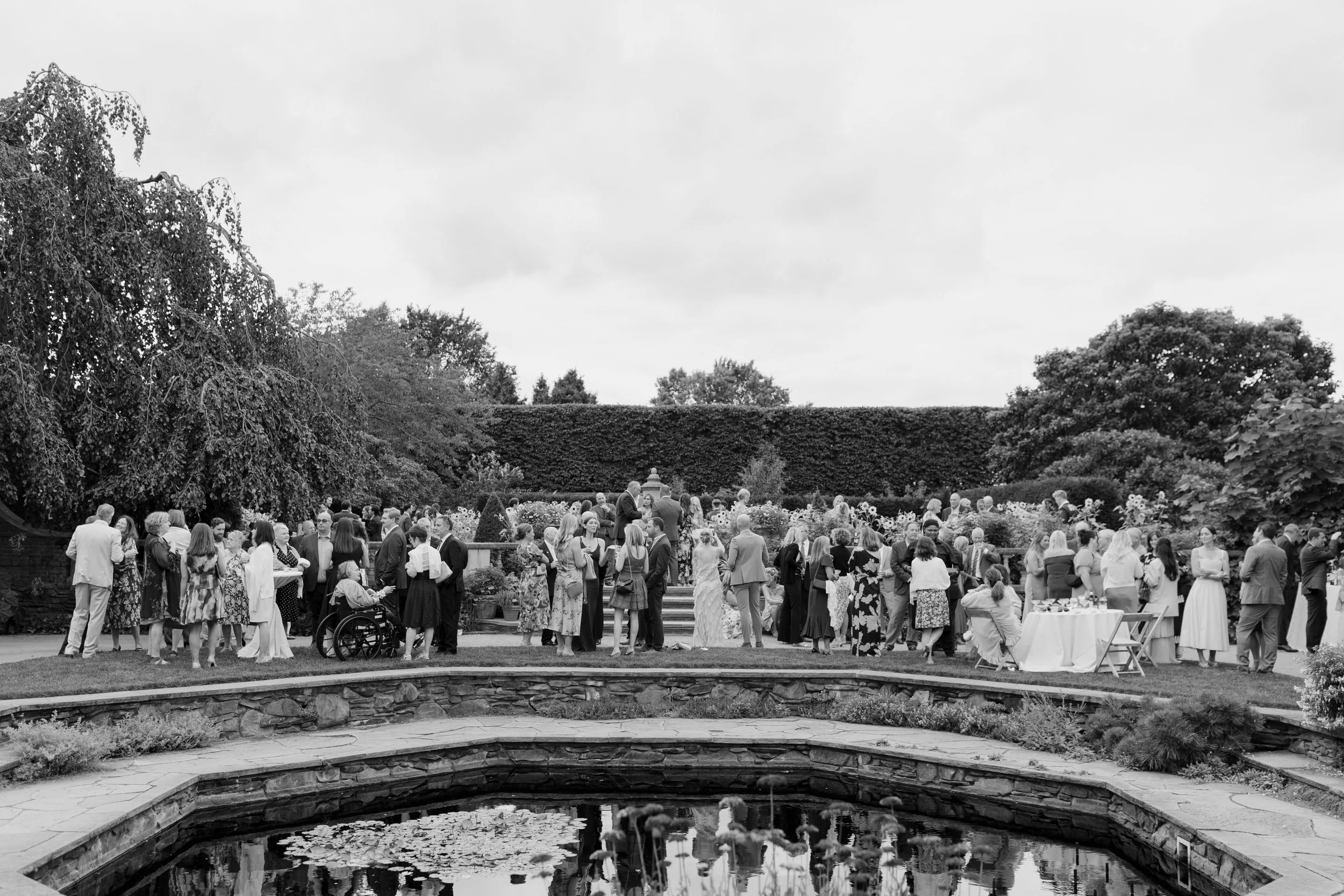 A large outdoor gathering of people at a garden party with tables, flowers, and a fountain in the foreground, trees, and a cloudy sky in the background.