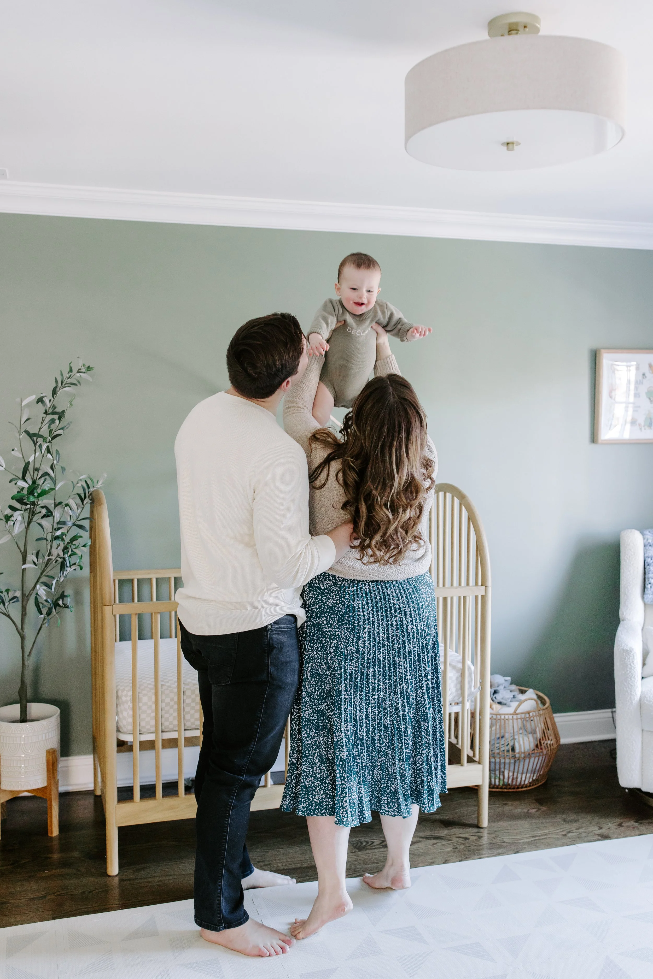A family holding a baby in a nursery room with green walls, a wooden crib, and a potted plant.