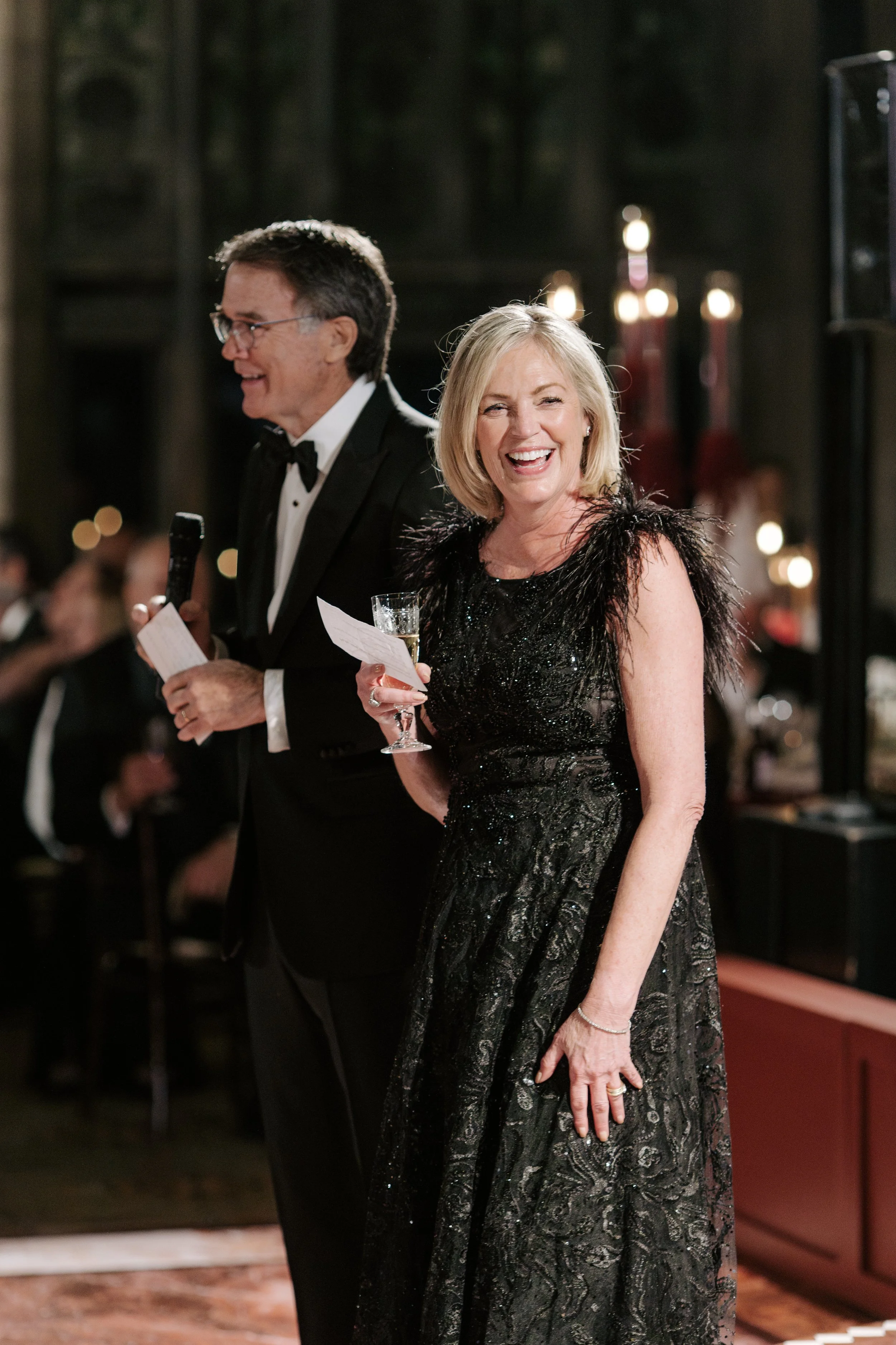 A man in a tuxedo holding a microphone and notes, and a woman in a black, shiny, beaded gown, both smiling and standing at an event with a dark background and blurred guests.