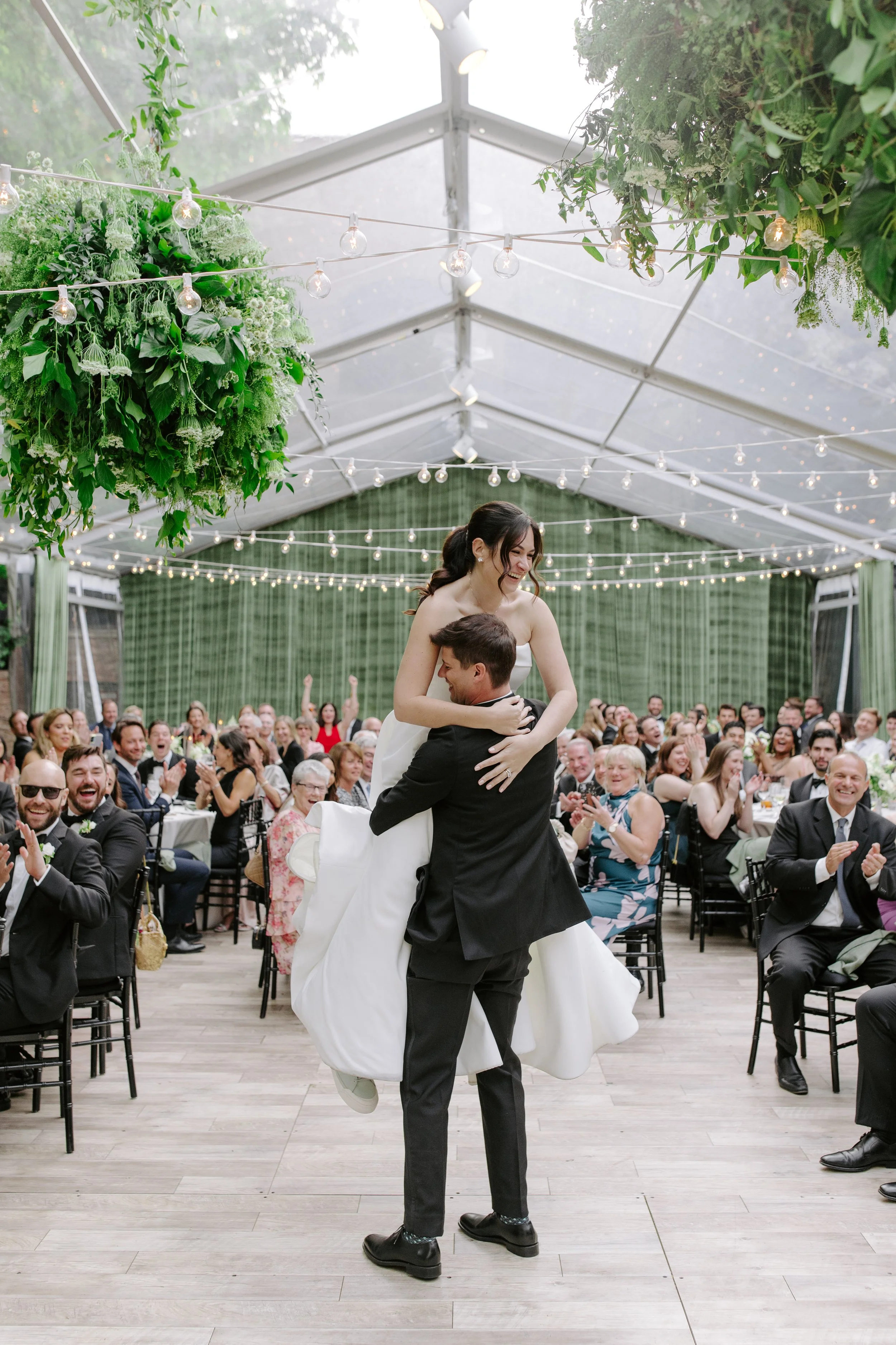 A bride and groom are dancing and being lifted by a guest at a wedding reception inside a tent decorated with string lights and hanging green floral arrangements. The bride is smiling and wearing a white wedding gown, while the groom is in a black tu