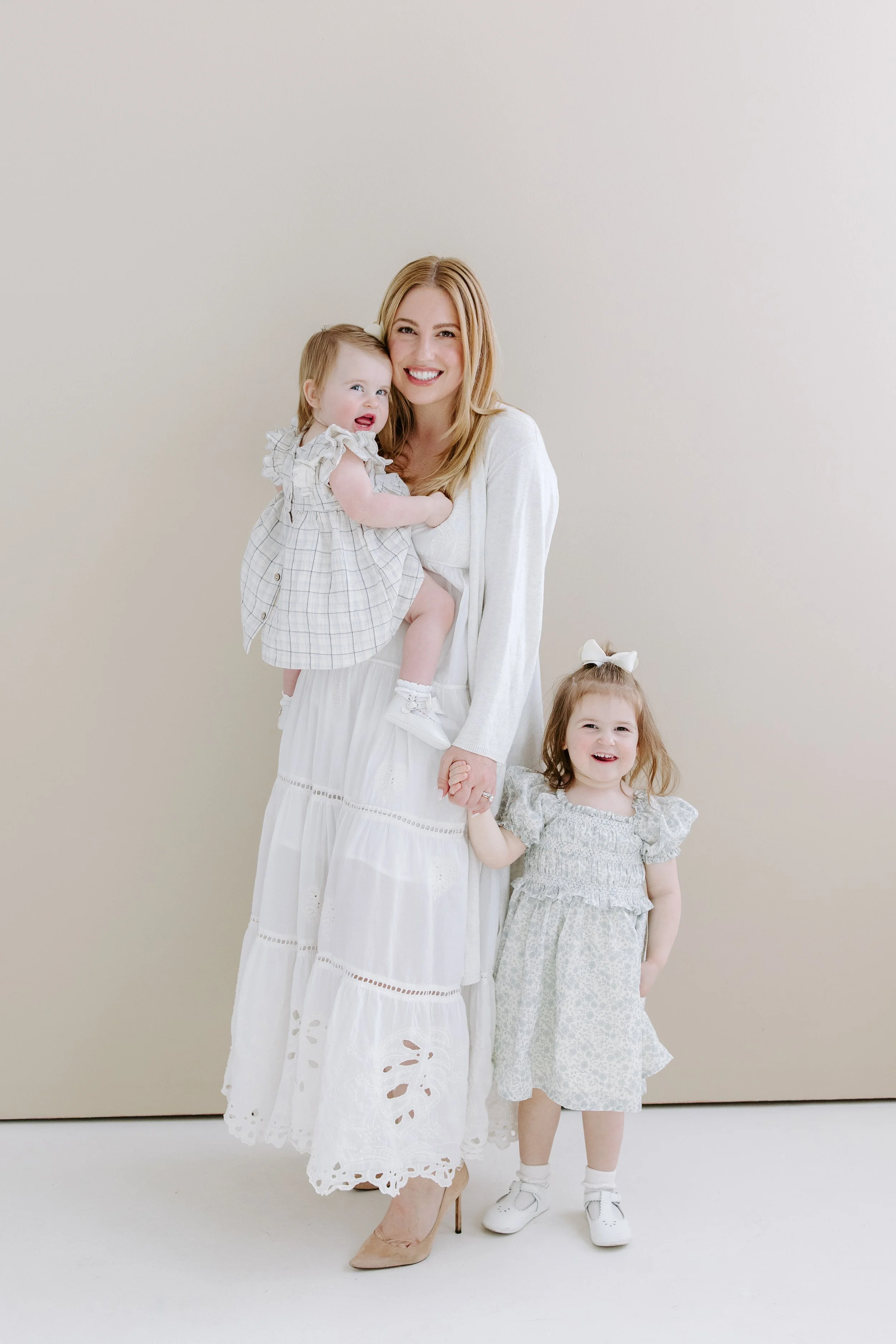 A woman with long blonde hair wearing a white dress and beige high heels is smiling while holding a young girl in her arms. The girl has light brown hair, is wearing a plaid dress with ruffled sleeves, and is making a playful expression. A second gir