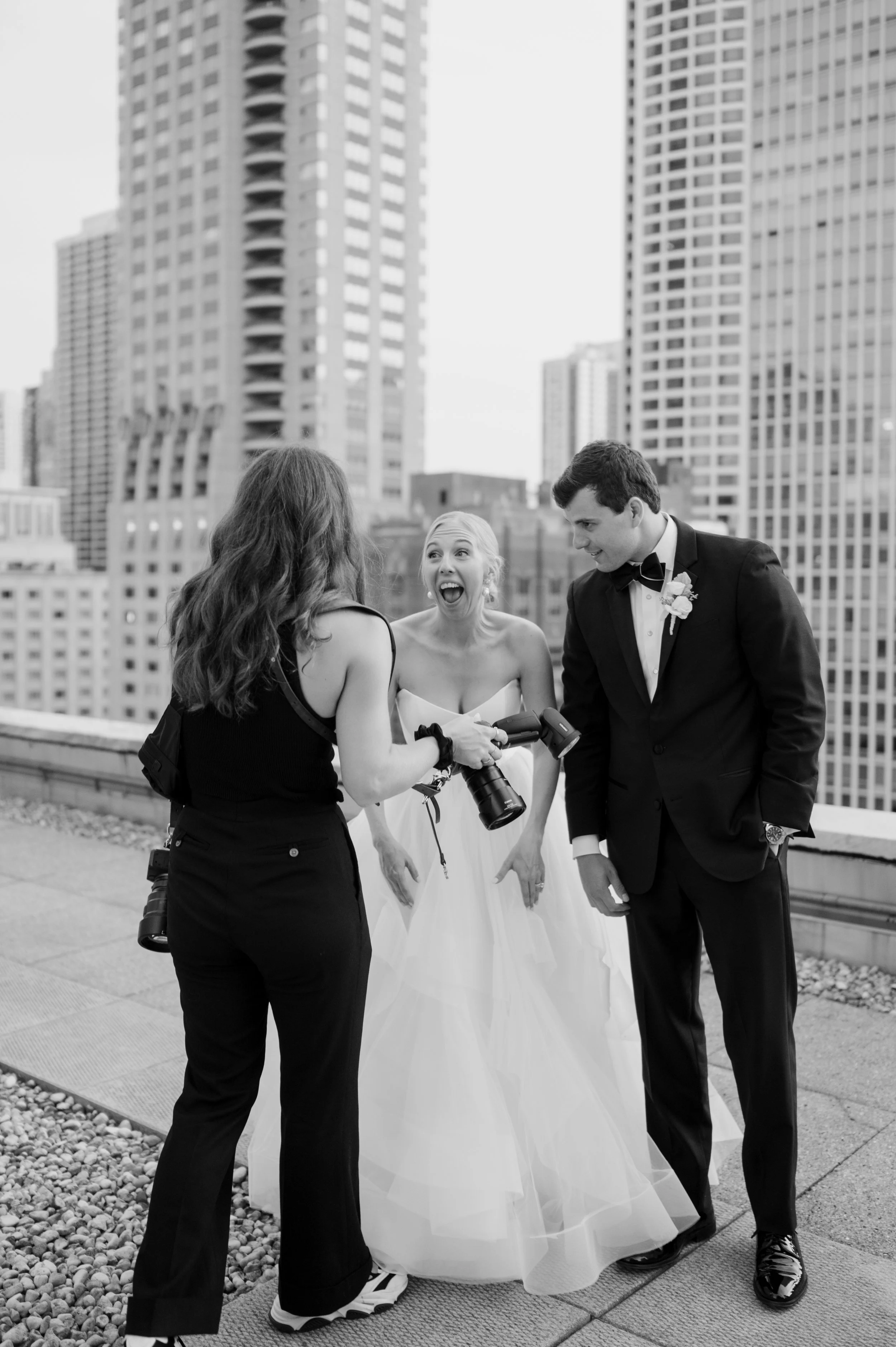 Black and white photo of a wedding scene on a rooftop with three people, a woman in a wedding dress, a man in a tuxedo, and a woman with long hair in casual clothing, all smiling and engaged in conversation with a city skyline of tall buildings in th