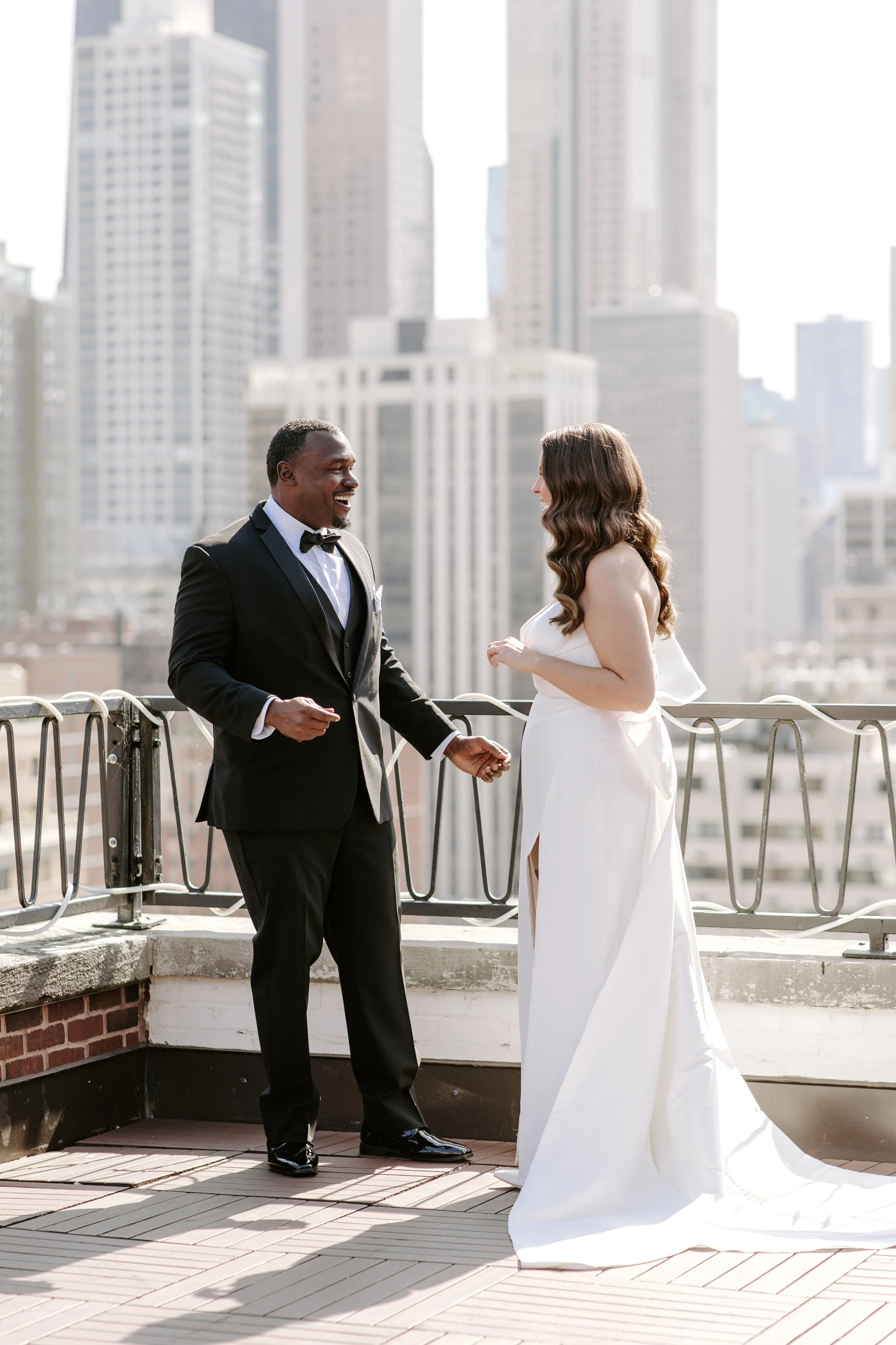 A couple in wedding attire on a rooftop, with city skyscrapers in the background. The groom is wearing a black tuxedo with a bow tie, and the bride is in a white wedding gown. They are holding hands and smiling at each other.