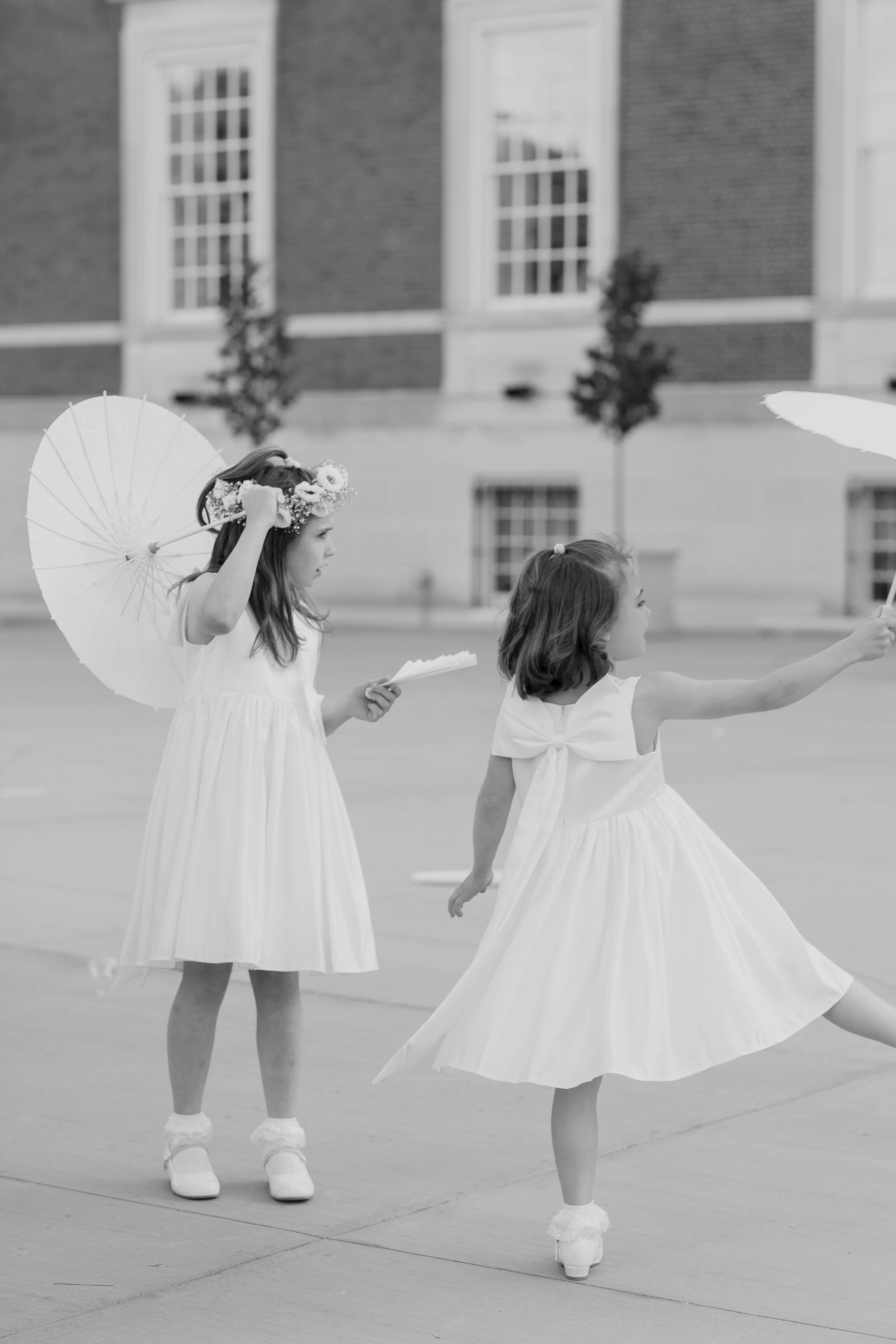 Two young girls in white dresses holding umbrellas and papers, participating in an outdoor event or celebration.