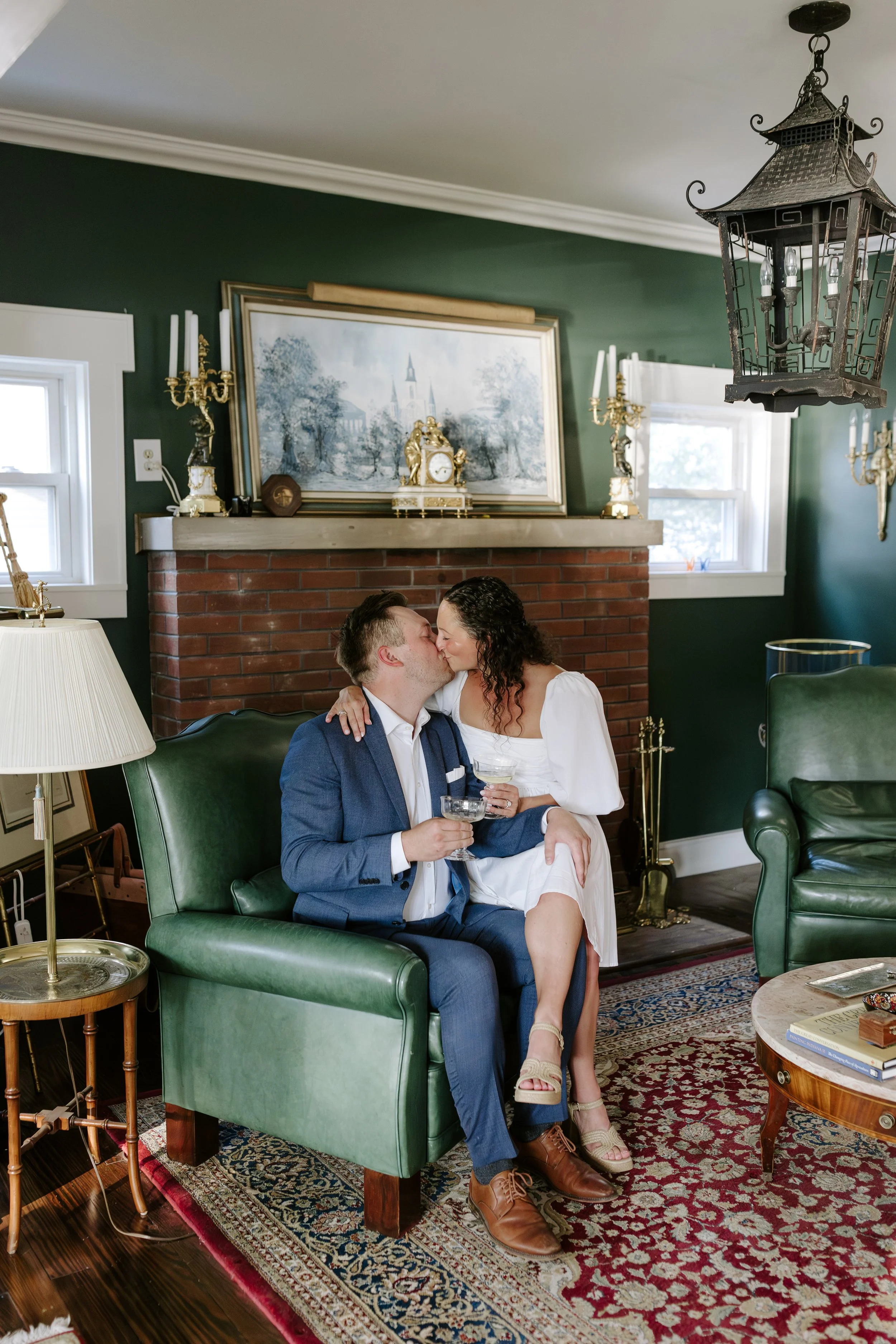 A man and a woman share a kiss on a green armchair in a decorated living room, holding champagne glasses. They are dressed semi-formally; the man wears a blue suit and brown shoes, and the woman wears a white dress and sandals. The room features a br