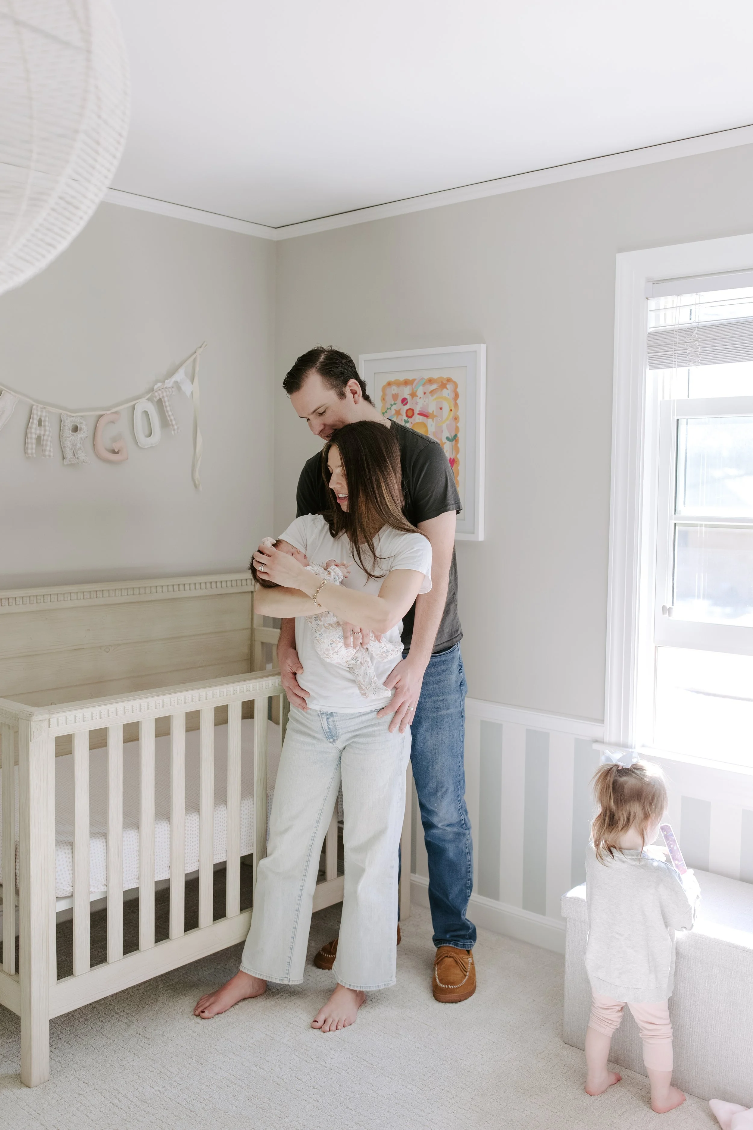 A couple holding a newborn baby in a softly lit nursery with a crib, wall art, and a window, two little girls in the room, one sitting on the floor and another near a box.