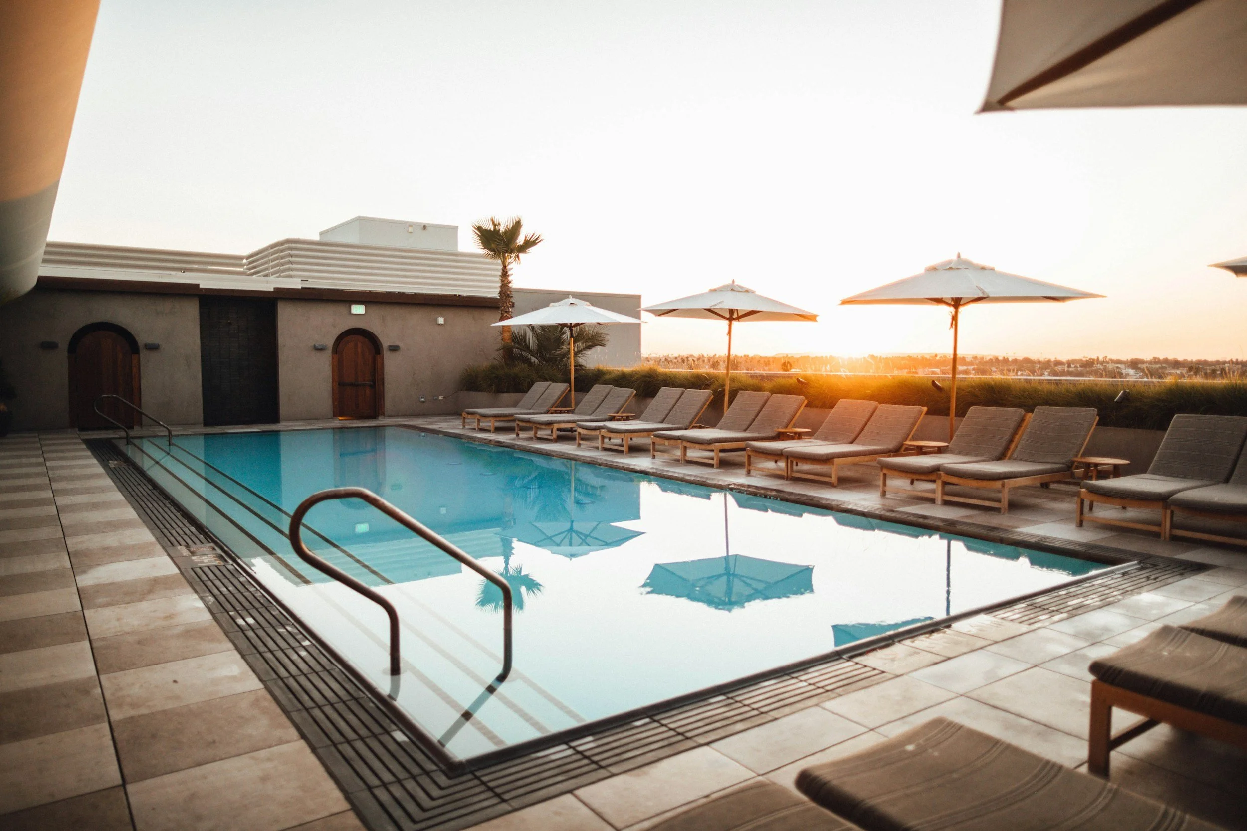 Sunset view of a hotel rooftop pool area with lounge chairs, umbrellas, a tall palm tree, and distant cityscape in the background.