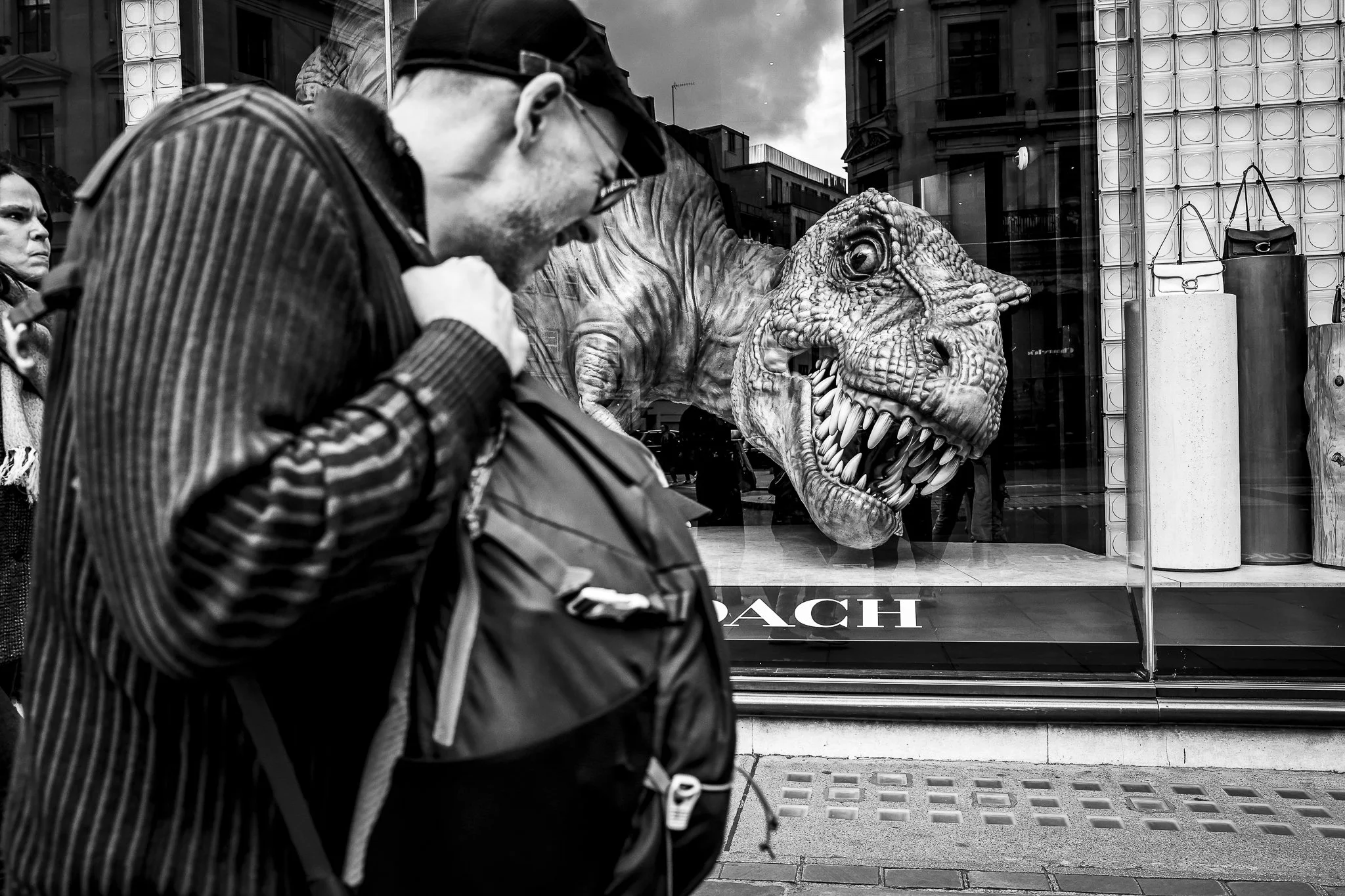 Black and white photo of two people walking past a storefront window with a large dinosaur head sculpture inside. One person, wearing glasses and a striped jacket, is in the foreground, and another person with a worried expression is also visible. Th