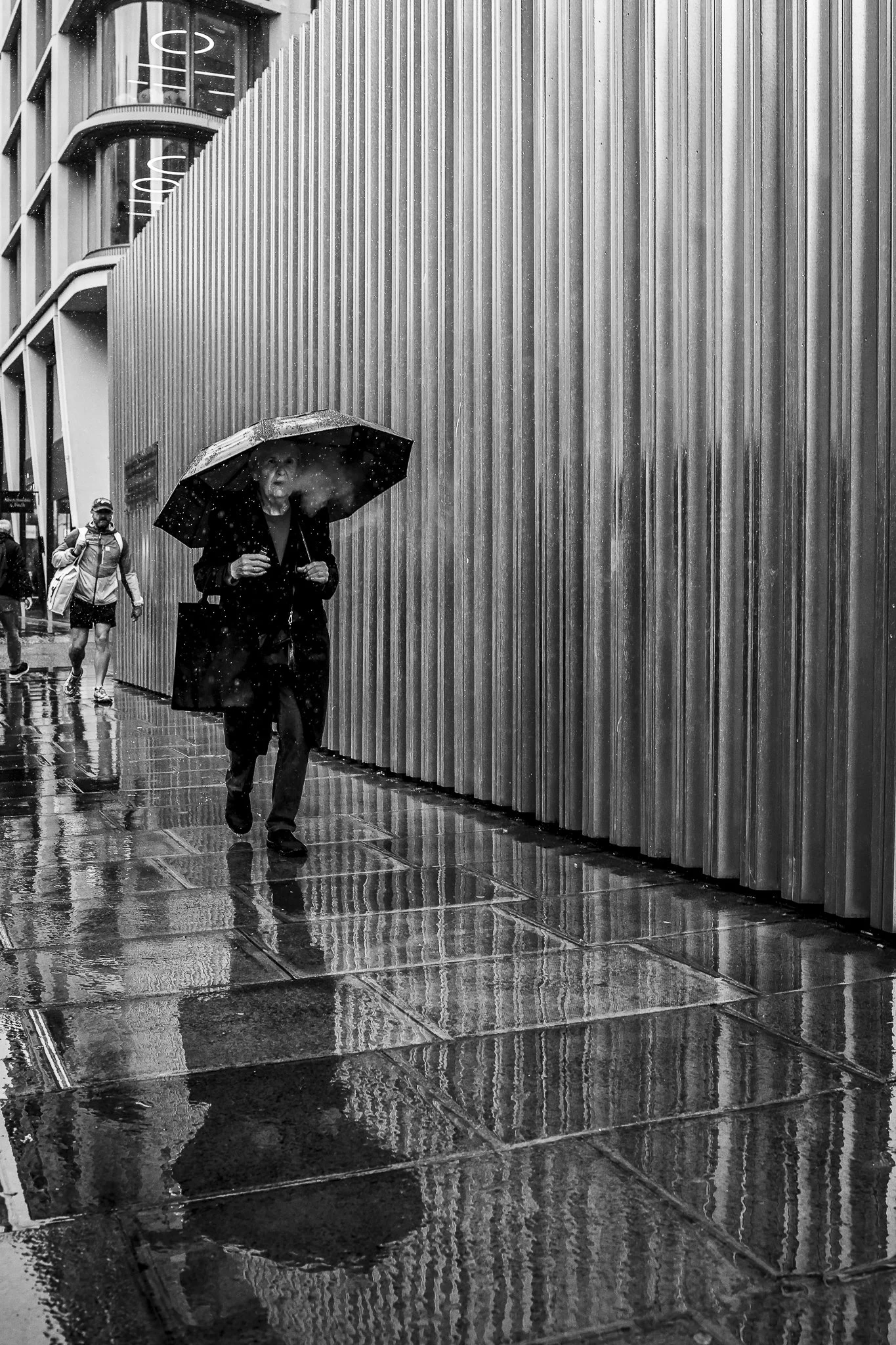 A woman walking with an umbrella on a rainy day, with reflections on the wet sidewalk and a modern building with vertical siding in the background.