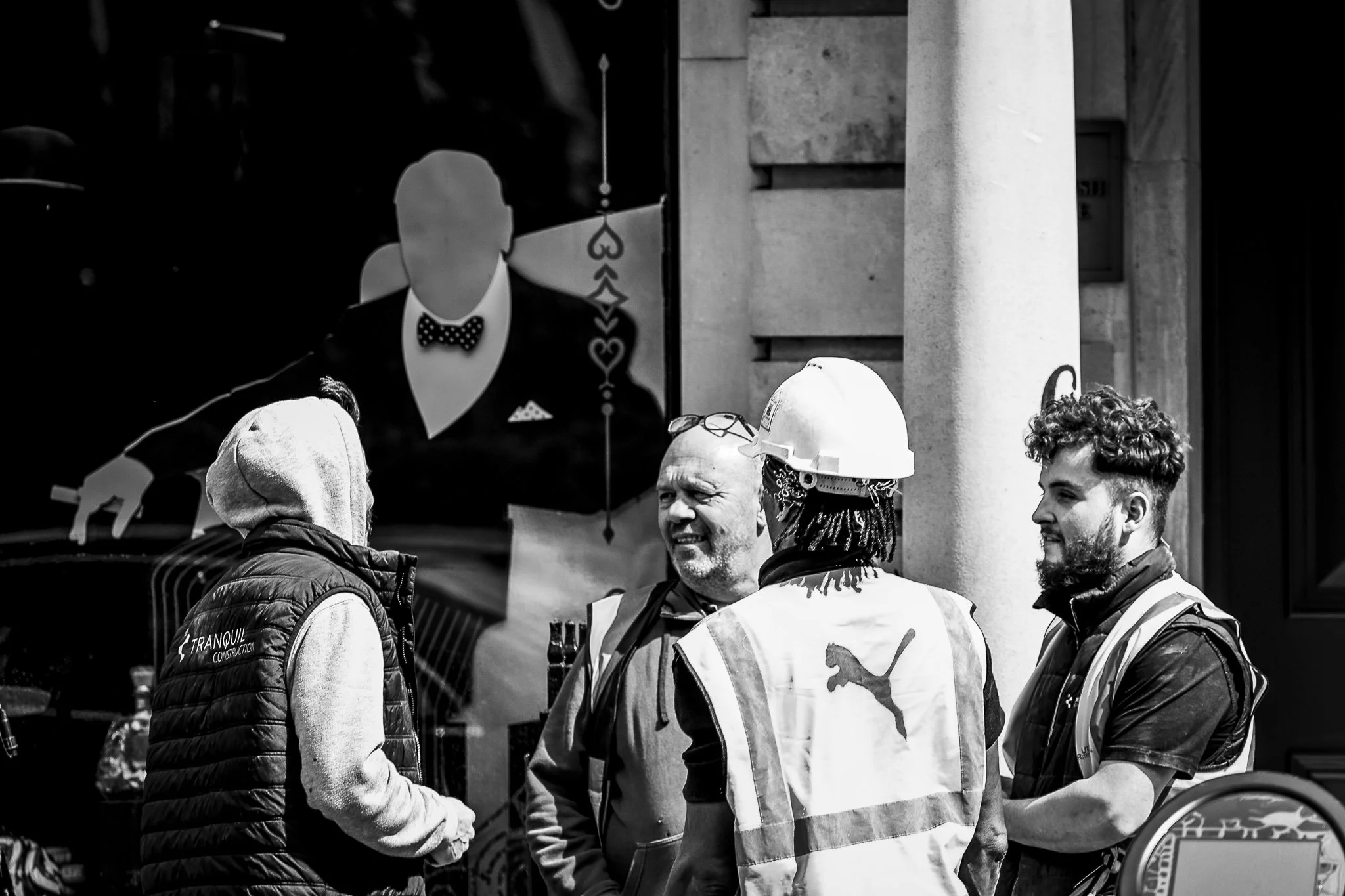 Group of people engaged in conversation on a city street, some wearing safety vests and hard hats, with a large sign featuring a stylized illustration of a woman in the background.