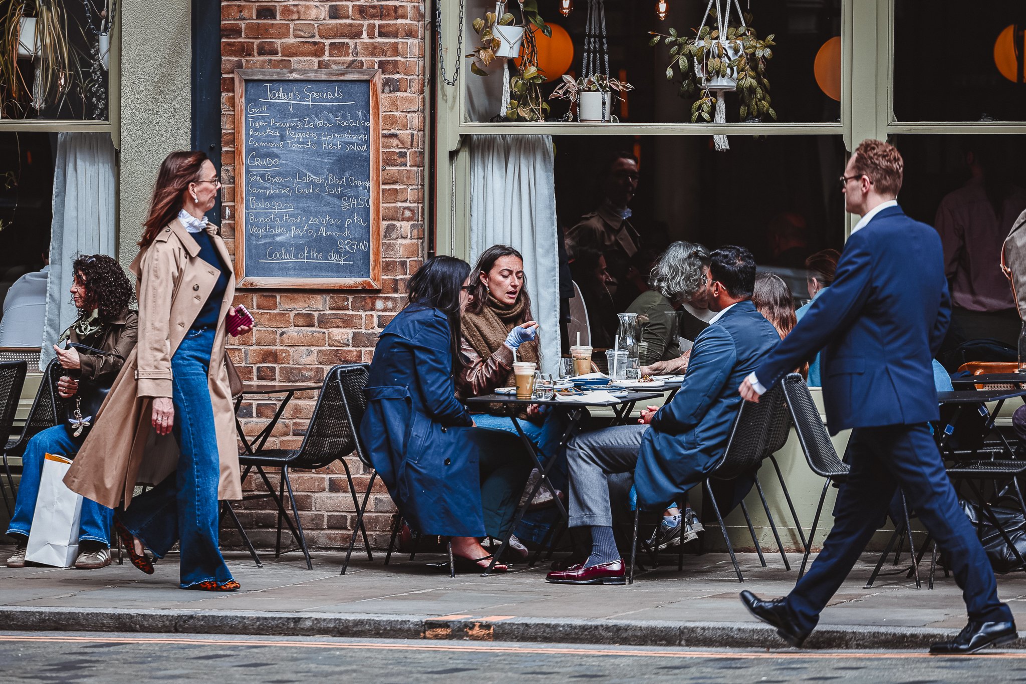 People sitting at tables outside a restaurant, with a chalkboard menu on the brick wall, and a woman walking past.