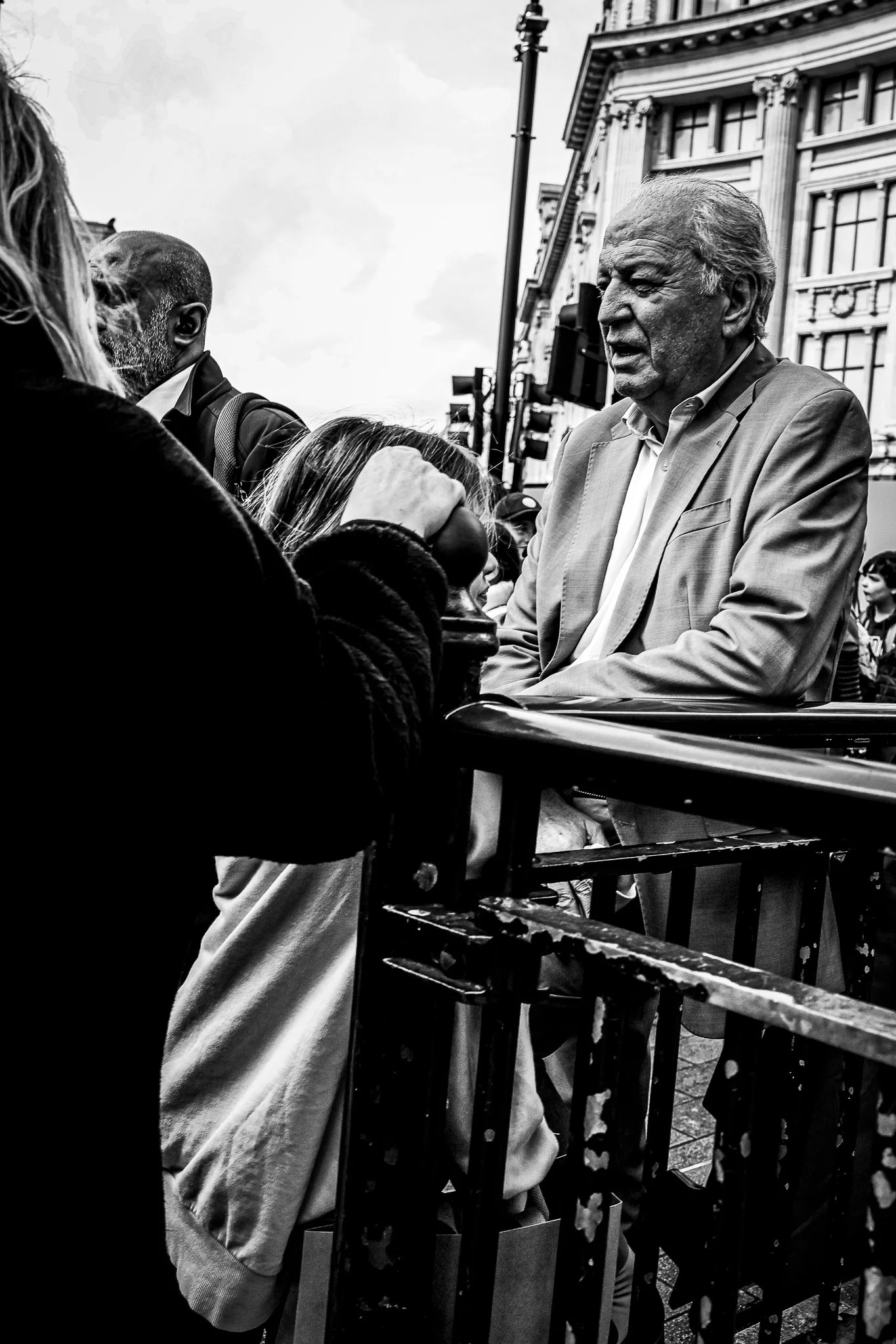 Black and white photo of a group of people, including an older man with gray hair and a suit, standing behind a metal railing on a city street with historic buildings in the background.