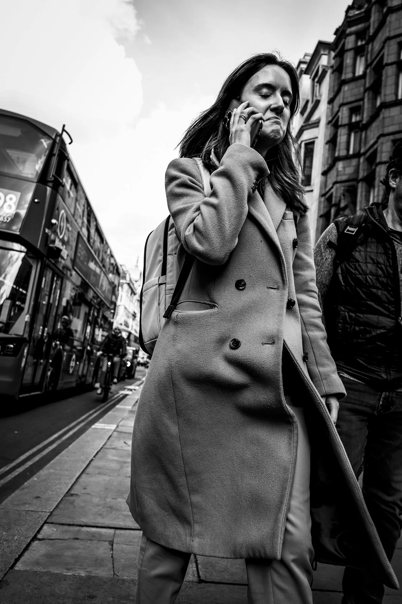 A woman with long hair, wearing a coat, is talking on a cell phone while walking on a city street. The background shows buildings and a double-decker bus.