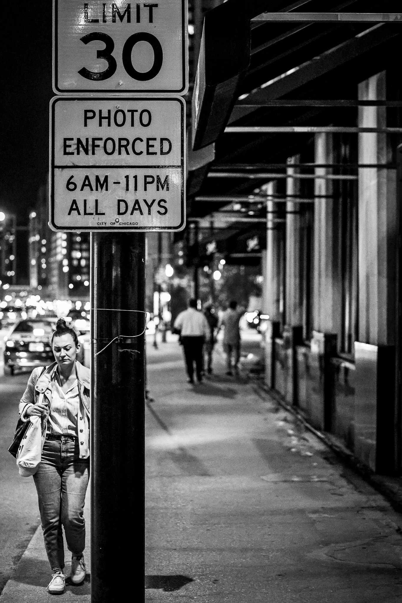 A black and white photo of a city street at night. A woman is walking near a sign that reads "Limit 30" and "Photo Enforced 6AM-11PM All Days." There are people and cars in the background, and a building with an awning on the right side of the image.
