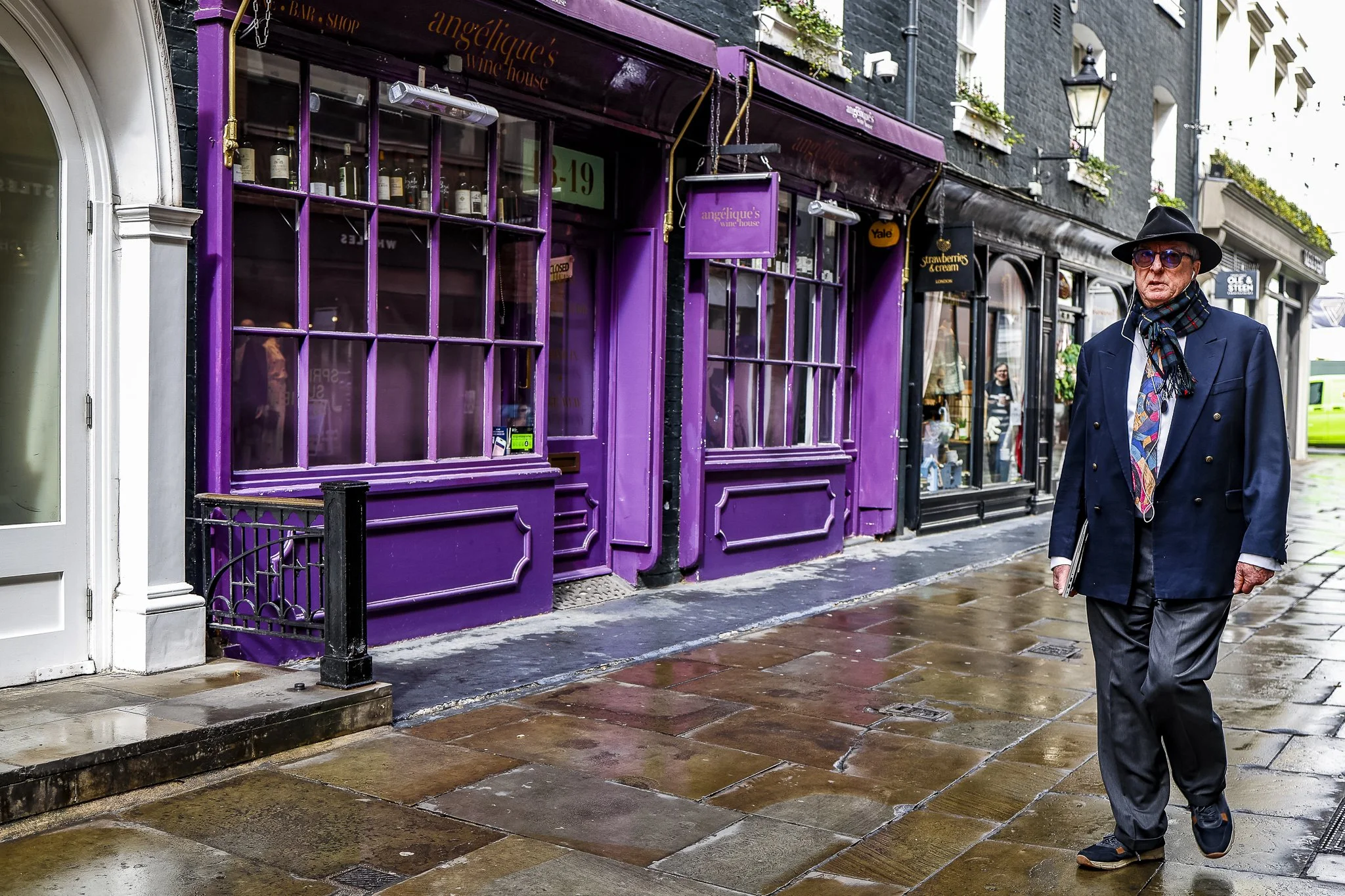 A city street with a purple storefront for 'Angelique's Wine House' and other shops. An older man wearing sunglasses, a dark hat, a blazer, and a scarf is walking on the wet sidewalk carrying a folder. The scene appears to be during or after rain.