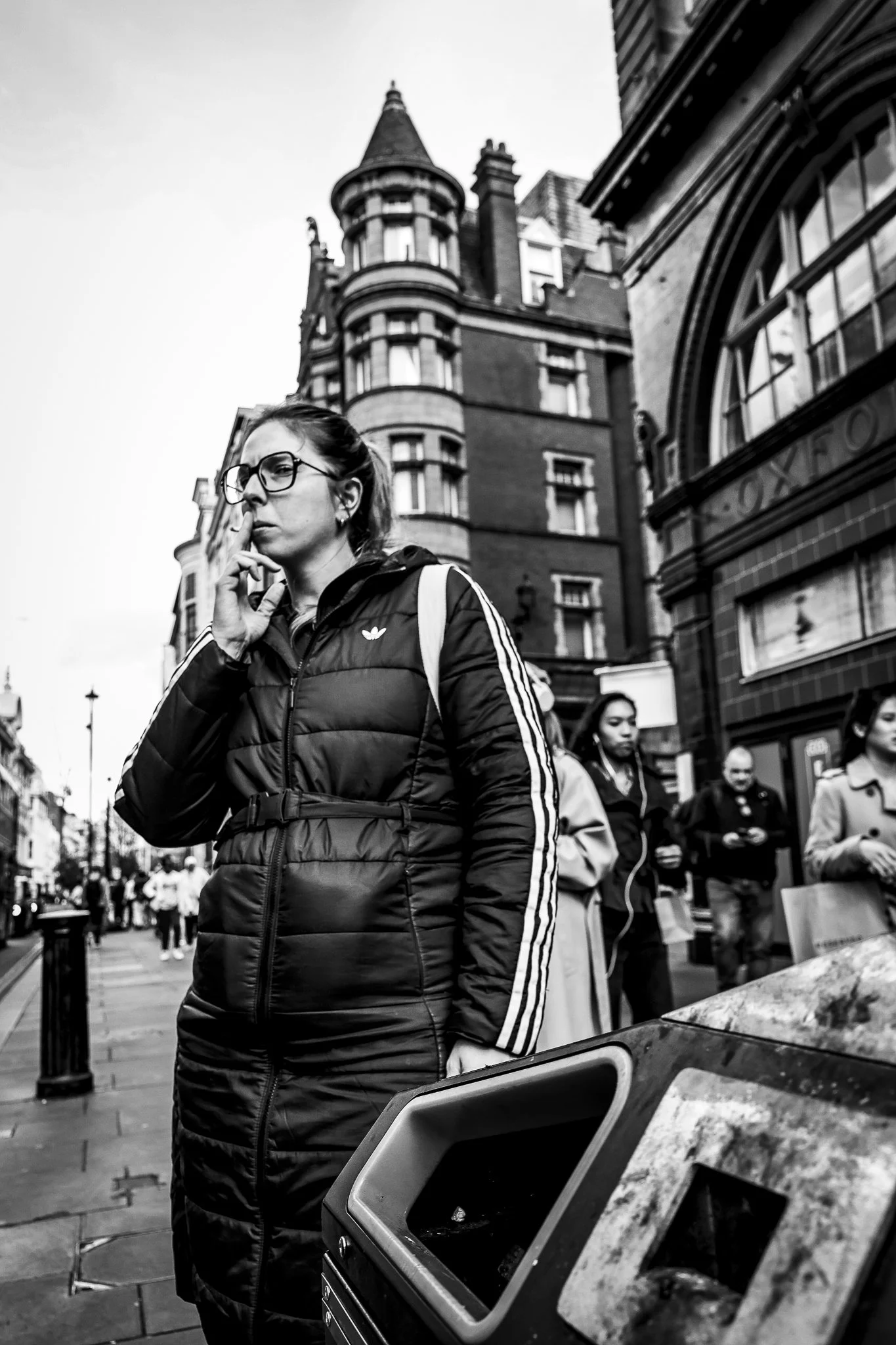 A woman with glasses and a puffy jacket standing on a city street with tall buildings in the background, a food truck in the foreground, and other people walking around.