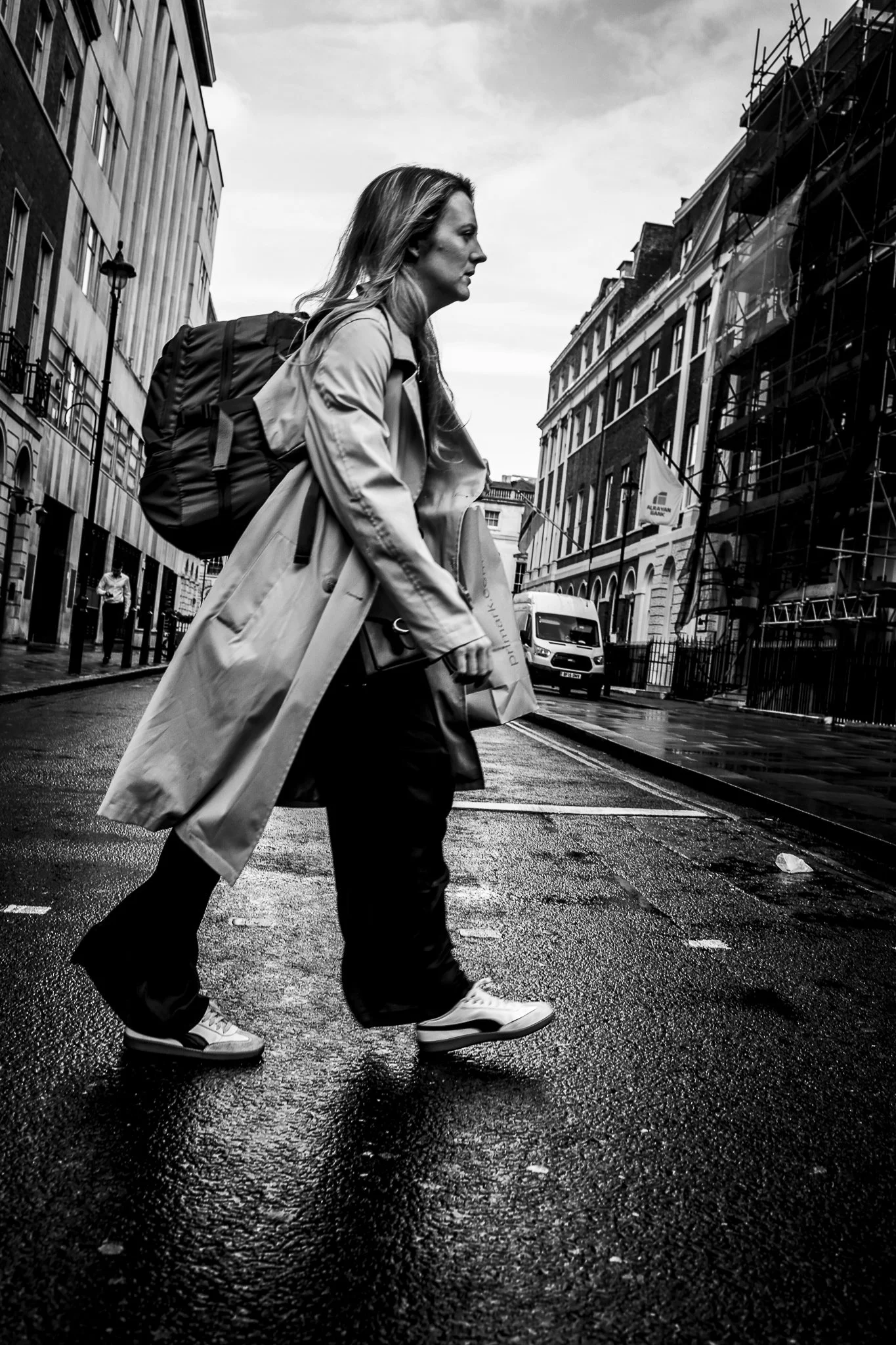 A woman with long hair carrying a backpack walking across a wet city street during overcast weather, with buildings and scaffolding in the background.