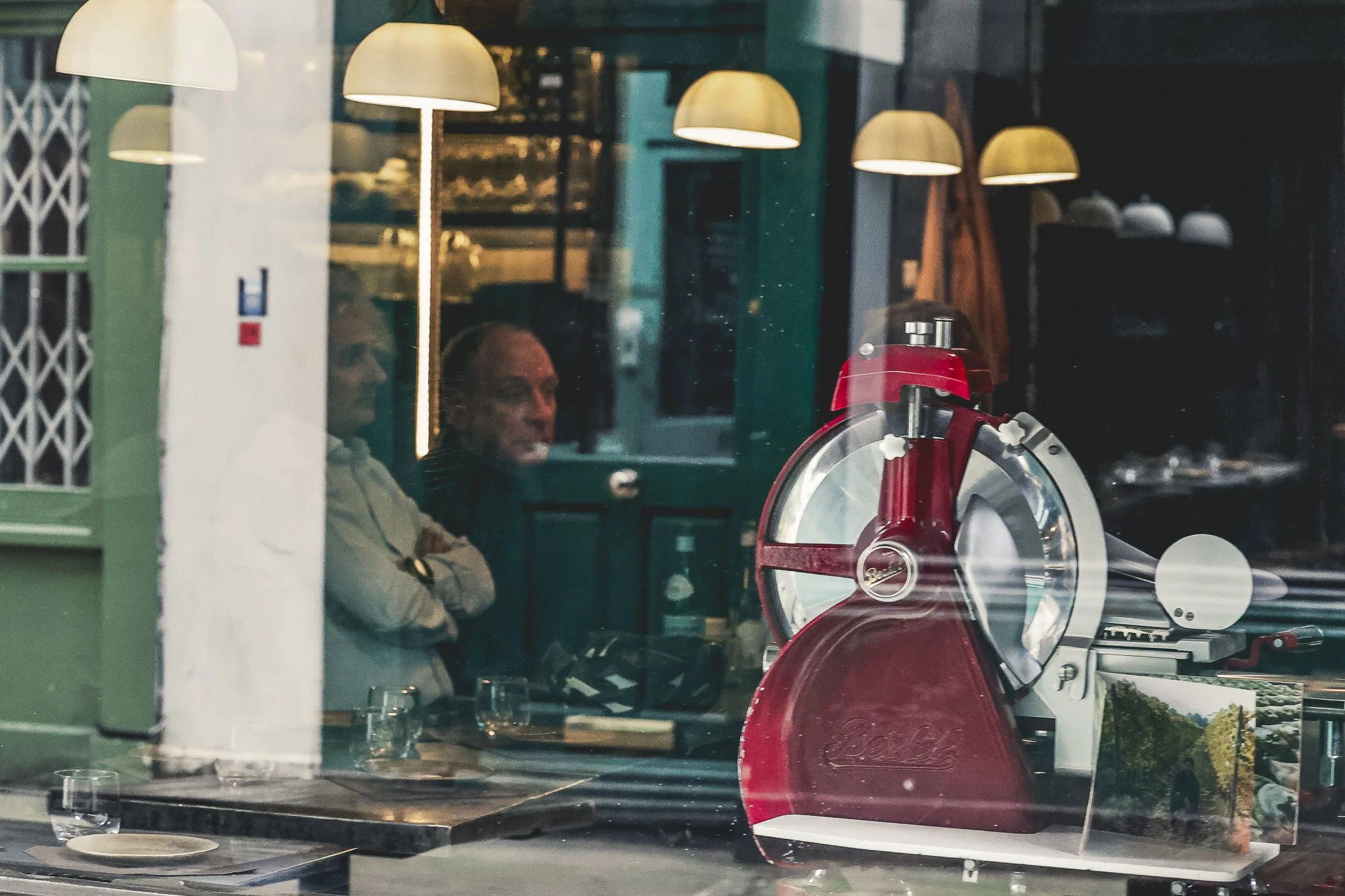 Inside a cafe or restaurant, with two people sitting at a table seen through a window. The reflection shows hanging lamps and wall decor, including a menu. There is a red pizza cutter on the table.