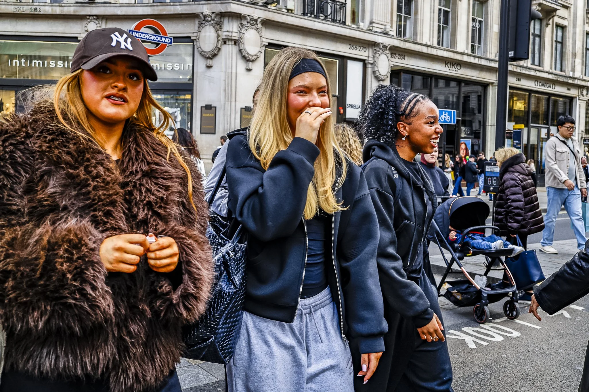 A group of diverse women standing and walking outdoors on a city street, some smiling and one covering her mouth in laughter. Background includes buildings, store signs, and pedestrians.