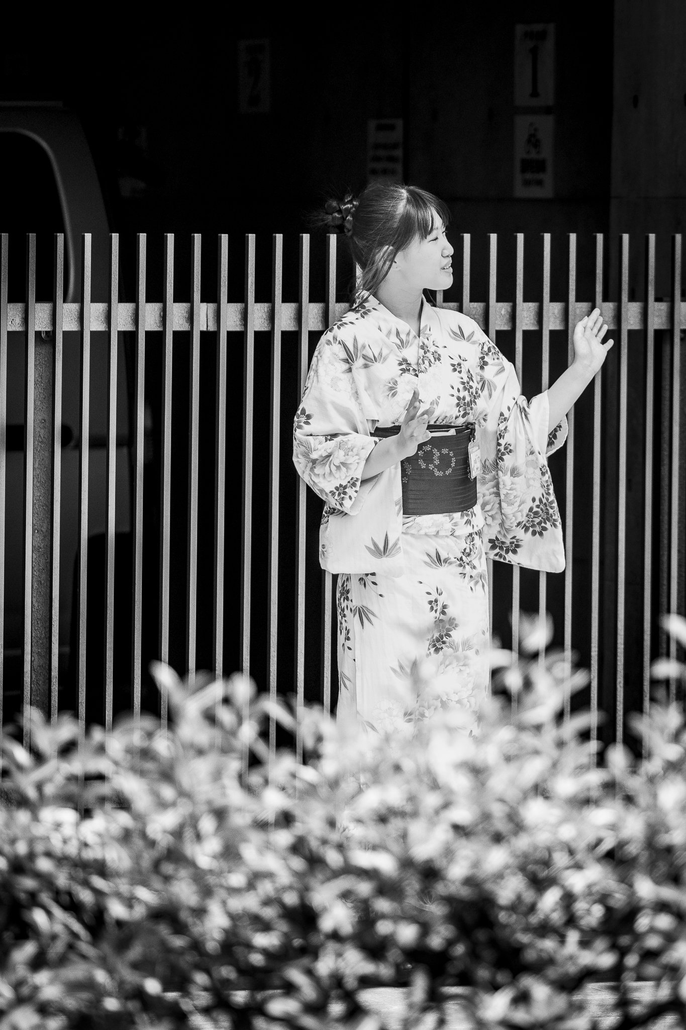A woman in a traditional kimono standing outside, smiling, and reaching out with her hand against a wooden fence, with a dark background.