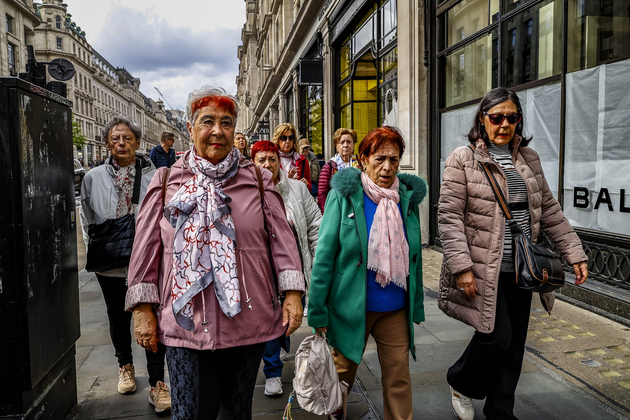 A group of older women walking on a city sidewalk, dressed in colorful jackets and scarves, with buildings and storefronts in the background.