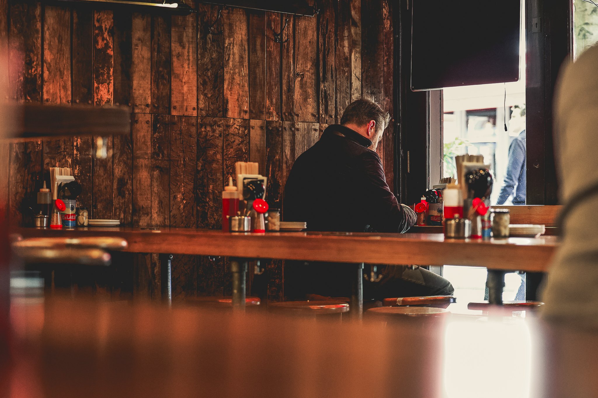 A person sitting alone at a restaurant table near a window with wooden walls in the background. The scene is warm with dim lighting.