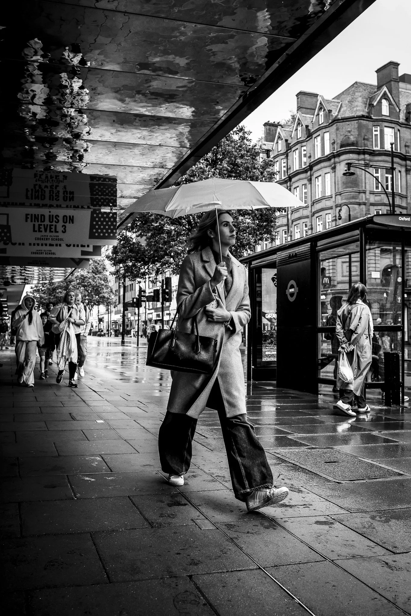 A woman walking on a rainy city street holding an umbrella, with buildings and other pedestrians visible in the background.