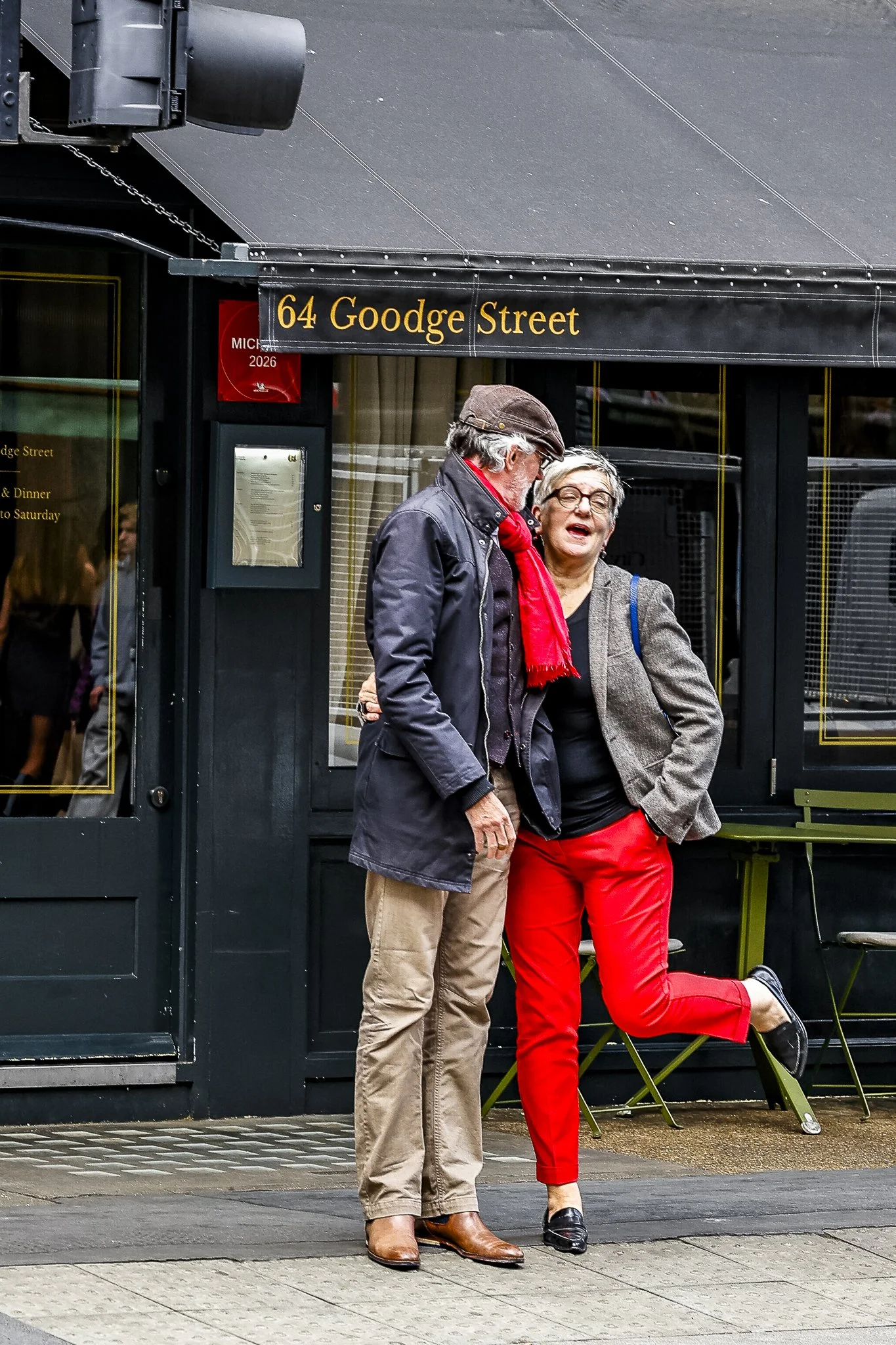 Two elderly people, a man and a woman, are standing outside near a storefront. The man is wearing a dark jacket, beige pants, and brown boots; the woman is dressed in a grey blazer, red pants, and black shoes, and she is raising one leg with a big sm