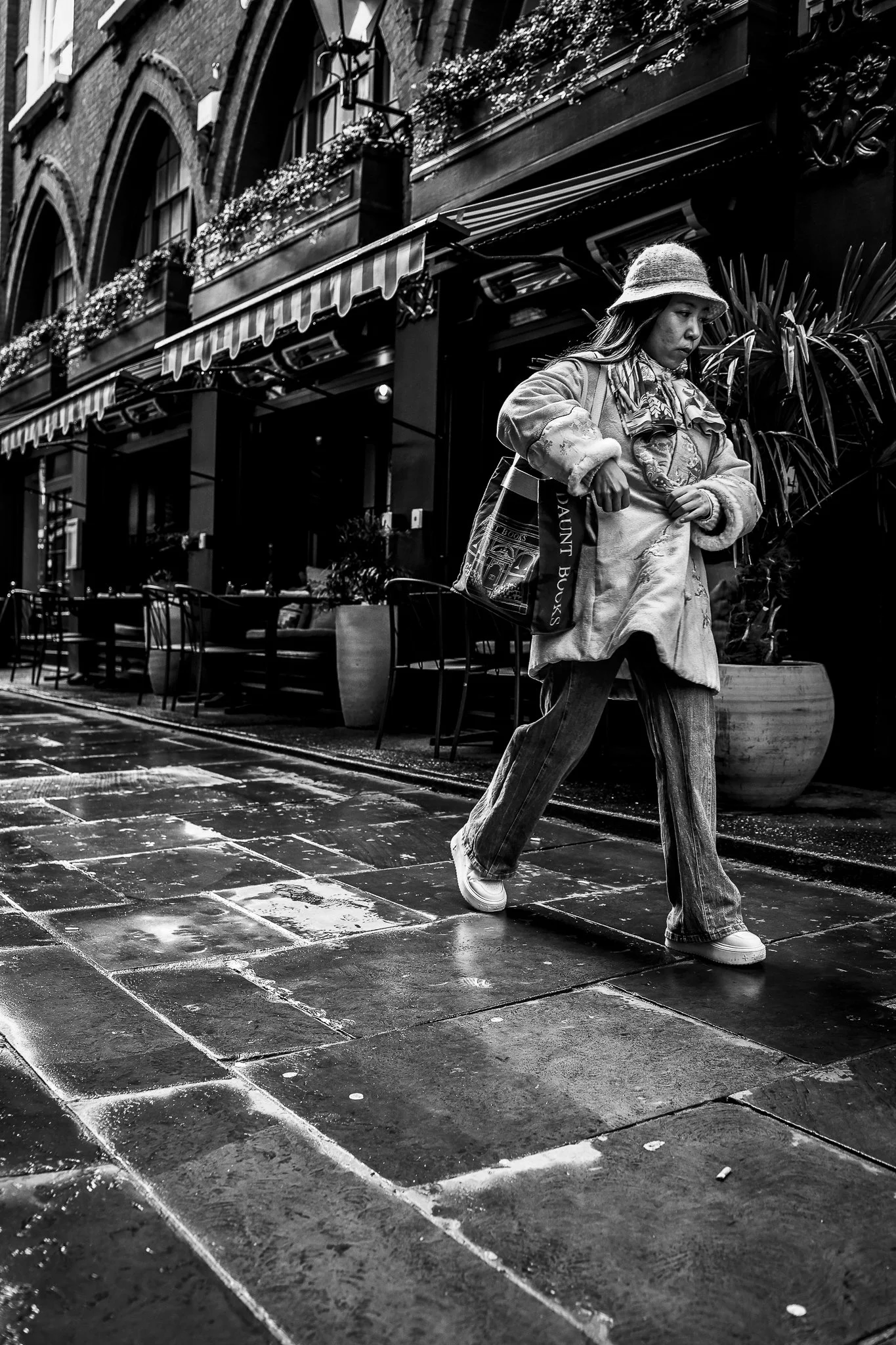 A woman walking on a wet city street with buildings and outdoor seating in the background, black and white photo.