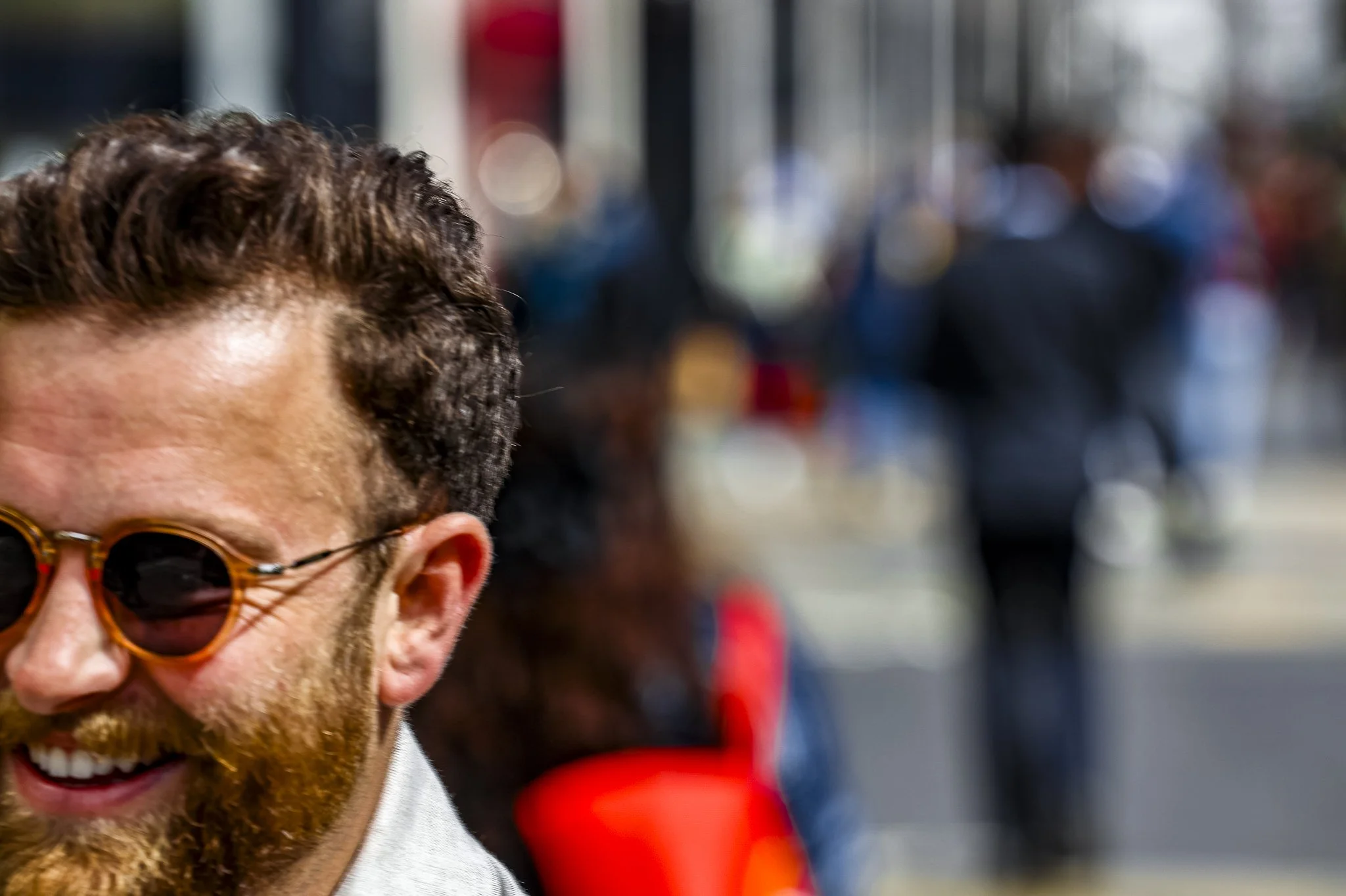 A man with curly hair and a beard, wearing sunglasses, is smiling in an outdoor, possibly urban setting.