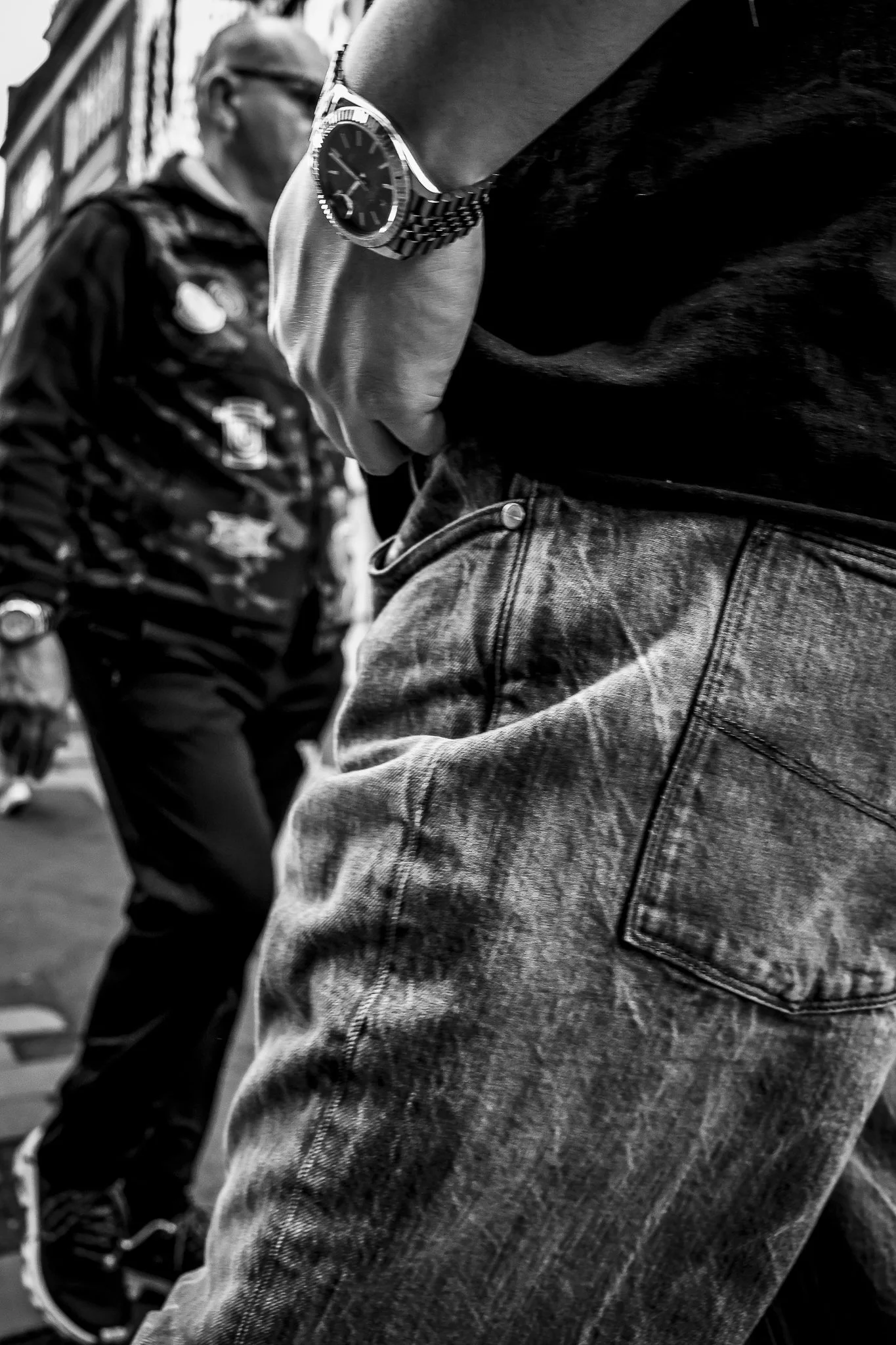 Close-up of a person's hand wearing a wristwatch, resting on their thigh, with another person in the background walking in an urban environment.