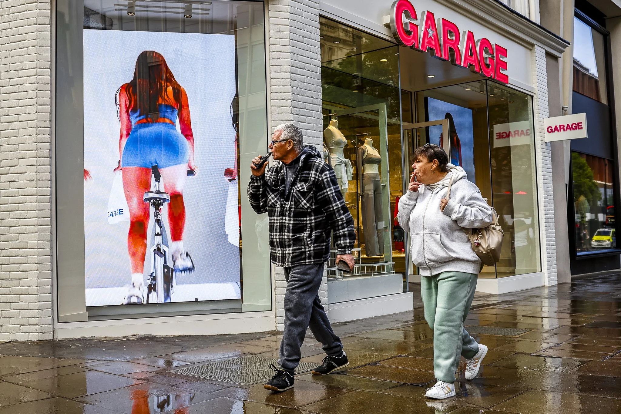 Two people walking past a store with a red sign that says "GARAGE" and a large digital billboard displaying a woman riding a bicycle, seen from behind, wearing a blue top and shorts. The scene takes place on a rainy day.