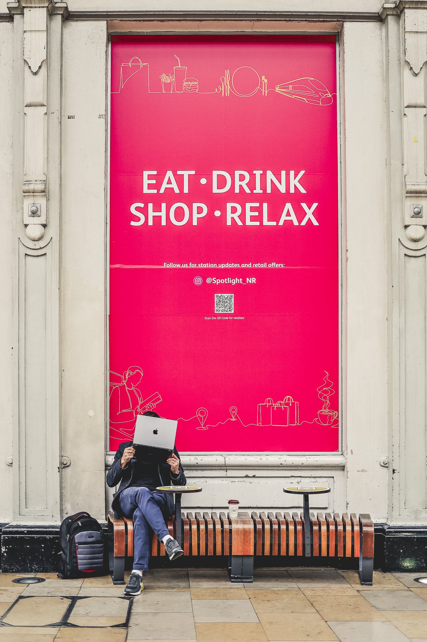 A man sitting on a bench with a laptop covering his face, holding it up. He has a backpack on the ground next to him and a cup on the bench. Behind him is a large pink advertisement featuring illustrations of a shopping bag, a drink, a burger, a plat