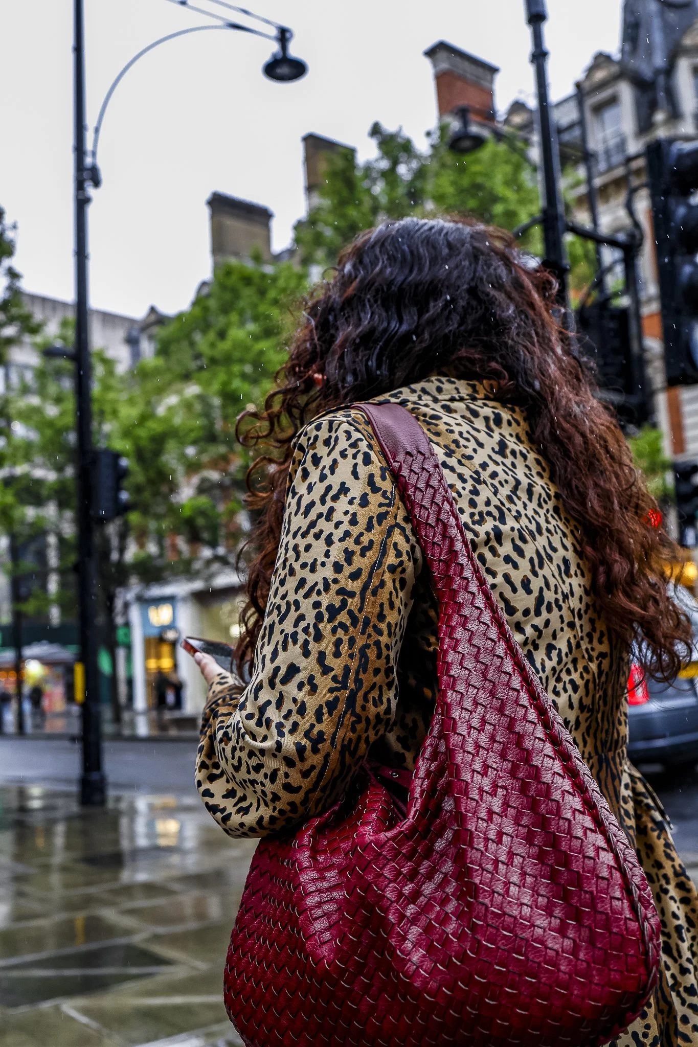A woman with long curly hair wearing a leopard print coat and carrying a large red woven leather bag, standing outdoors on a rainy day in an urban setting with buildings, street lamps, and trees in the background.
