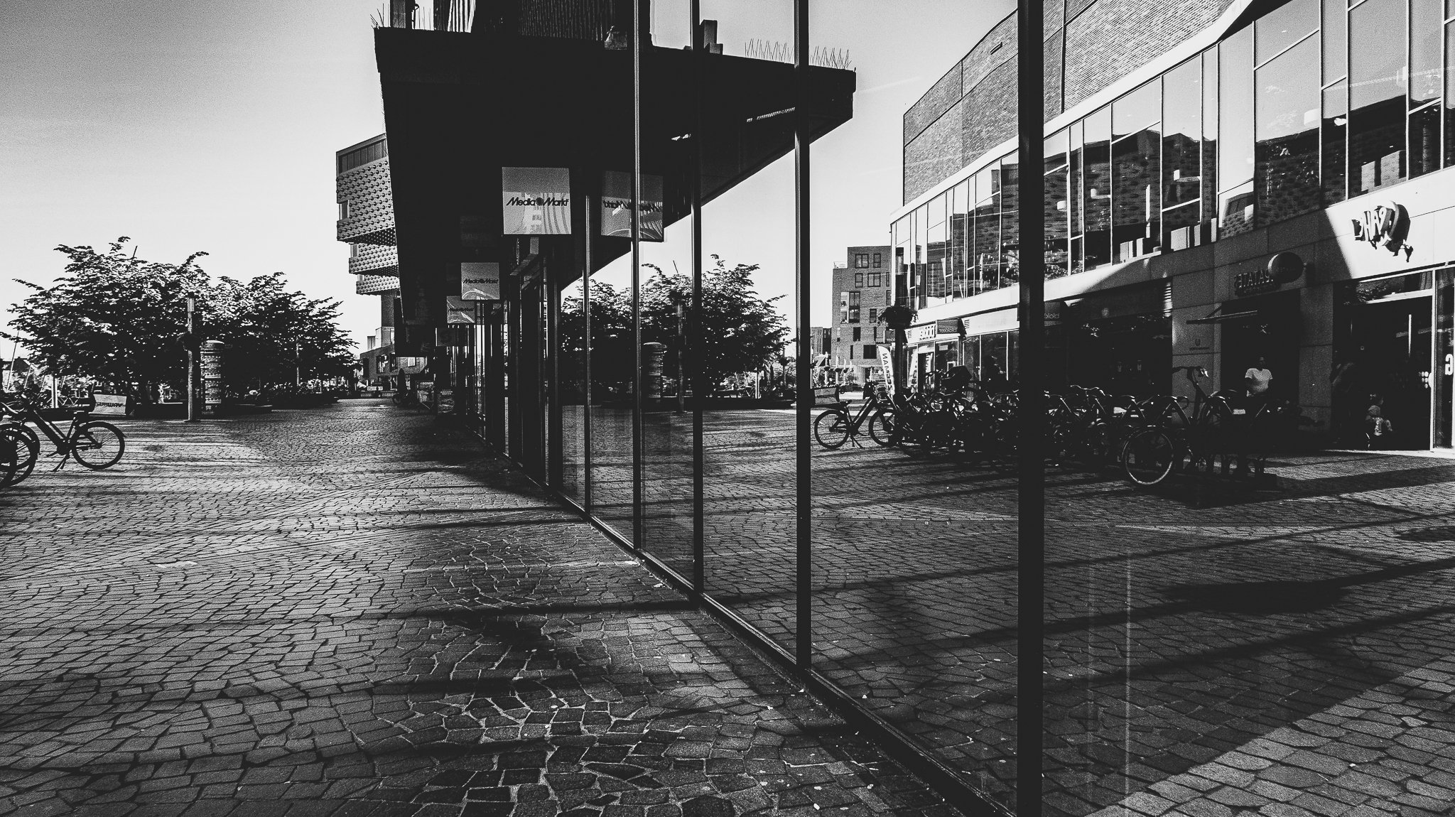 A city street scene in black and white showing modern buildings, bicycles parked on the sidewalk, trees, and a glass storefront reflecting the surroundings.