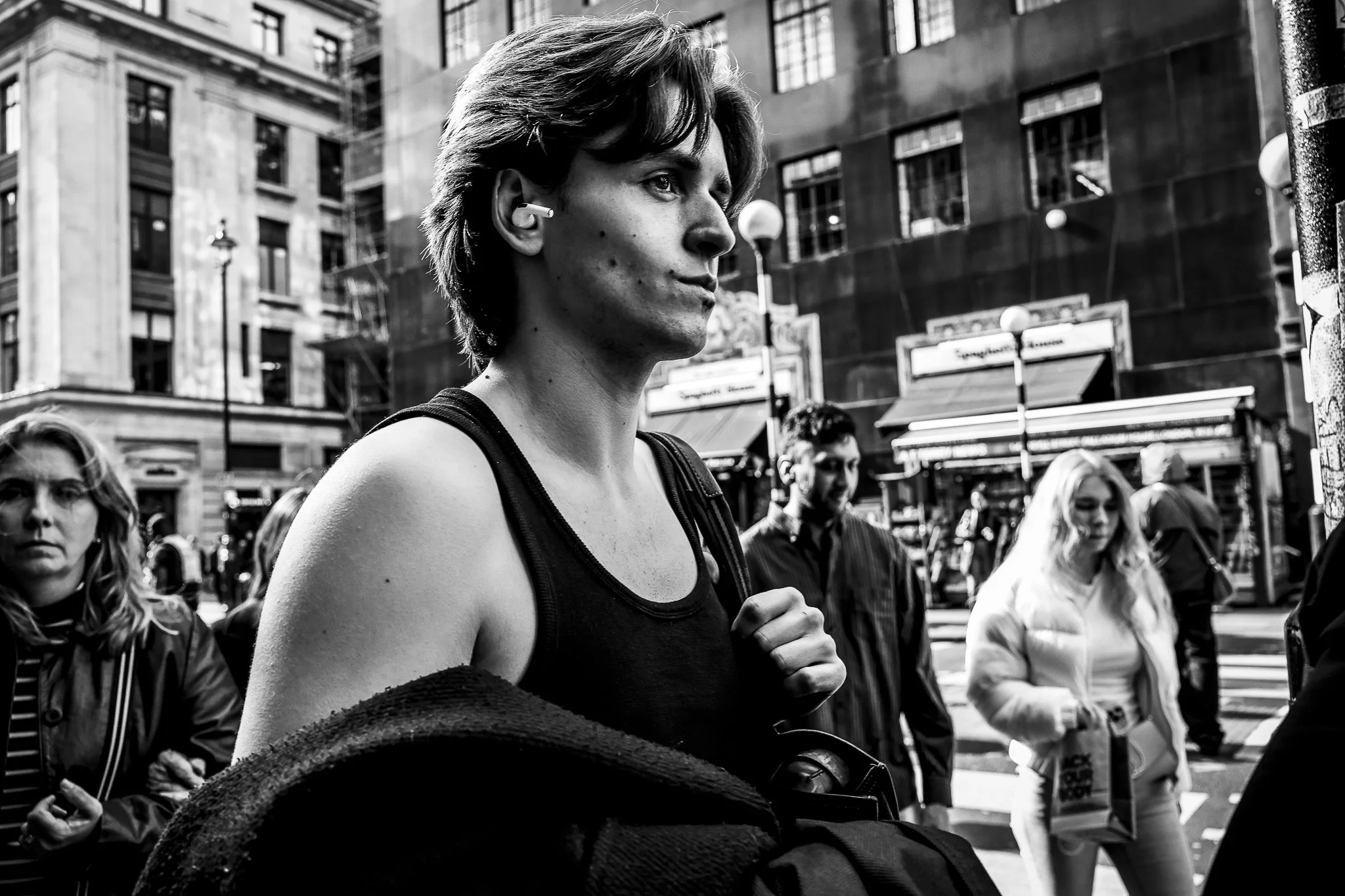 A woman with short hair wearing a tank top and wireless earbuds walking in a city with large buildings and street lamps, surrounded by other pedestrians.