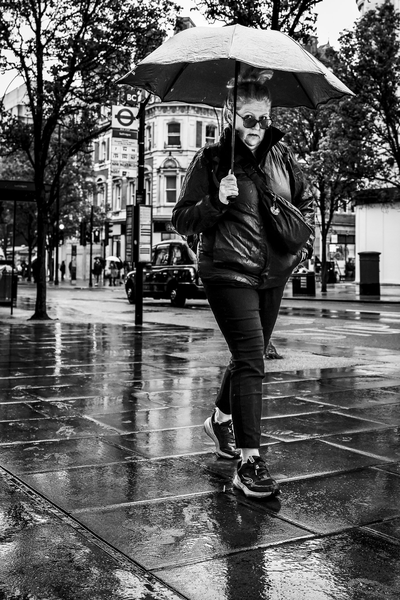 A woman walking on a wet city street with an umbrella, surrounded by trees and buildings in the background, captured on a rainy day.