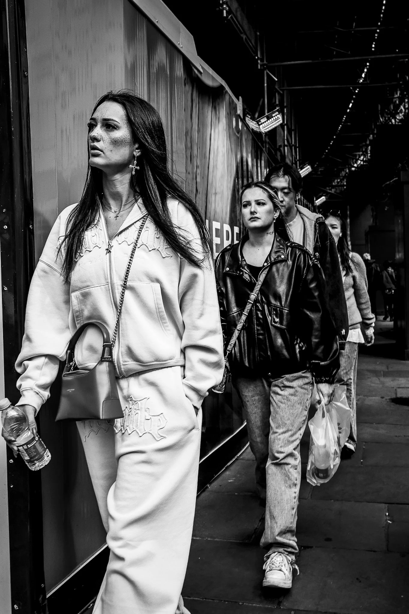 A black and white photo of a group of people walking in a line, some carrying shopping bags and one holding a water bottle, in an outdoor urban setting with metal structures and lights overhead.