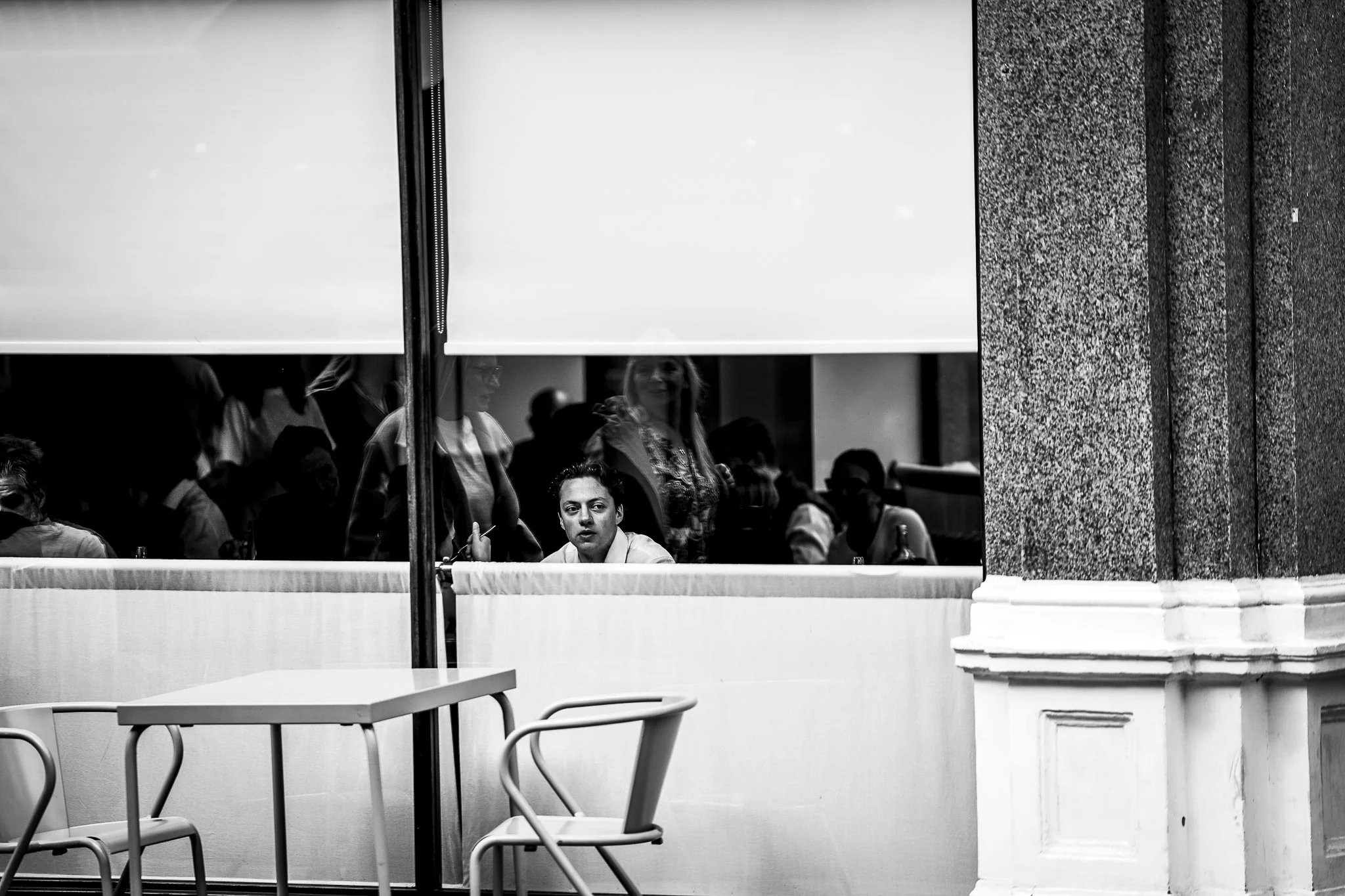 Black and white photo of people sitting at a counter inside a building, viewed through a glass window with blinds. Some people are visible inside, including a woman with long hair and a man with a serious expression, and others are reflected on the g