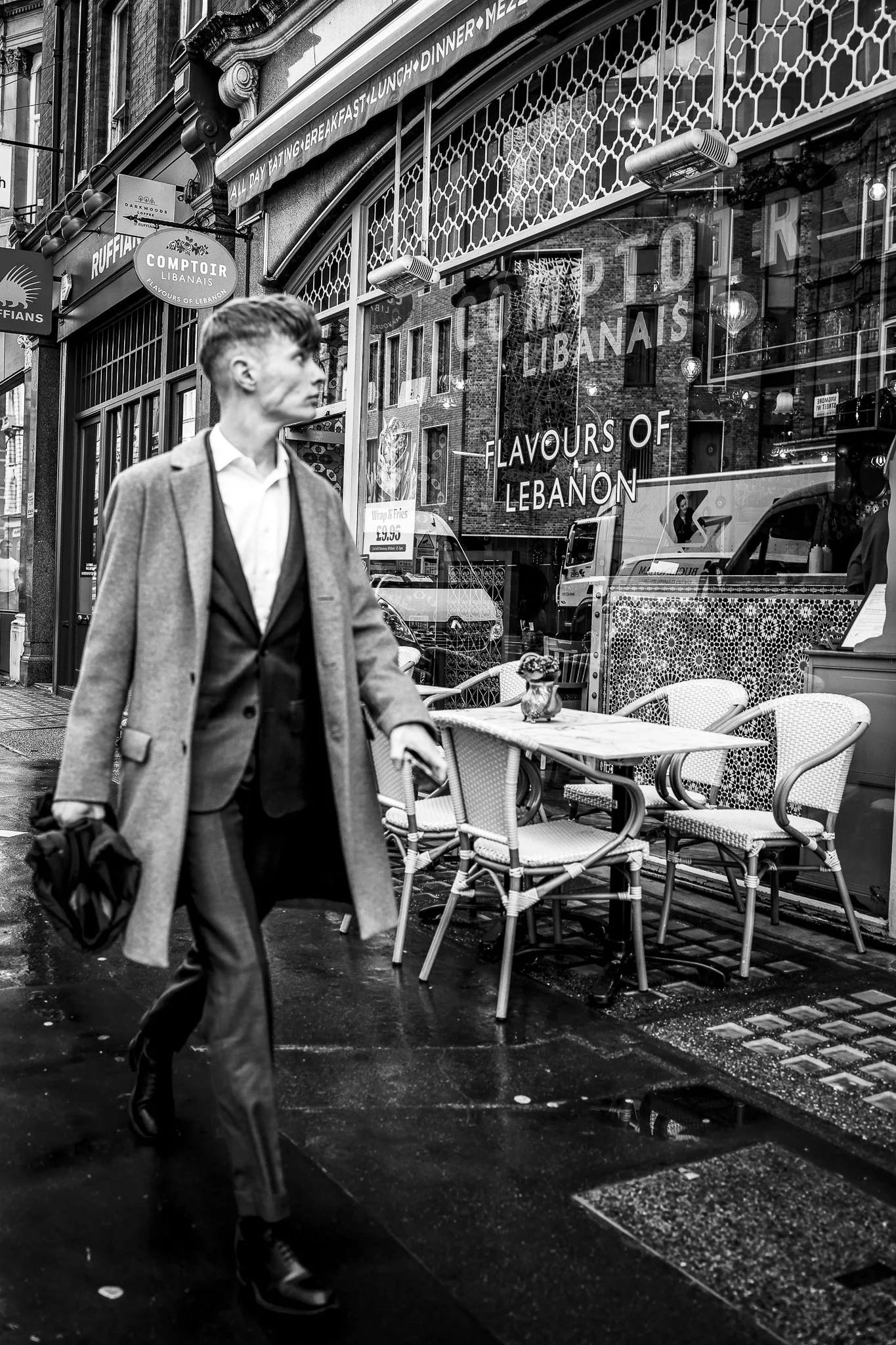 A black and white photo of a young man walking on a city sidewalk in front of a Lebanese restaurant with outdoor seating. The man is wearing a coat and seems to be holding a bag. The restaurant window displays signs for Lebanese flavors and a food pr