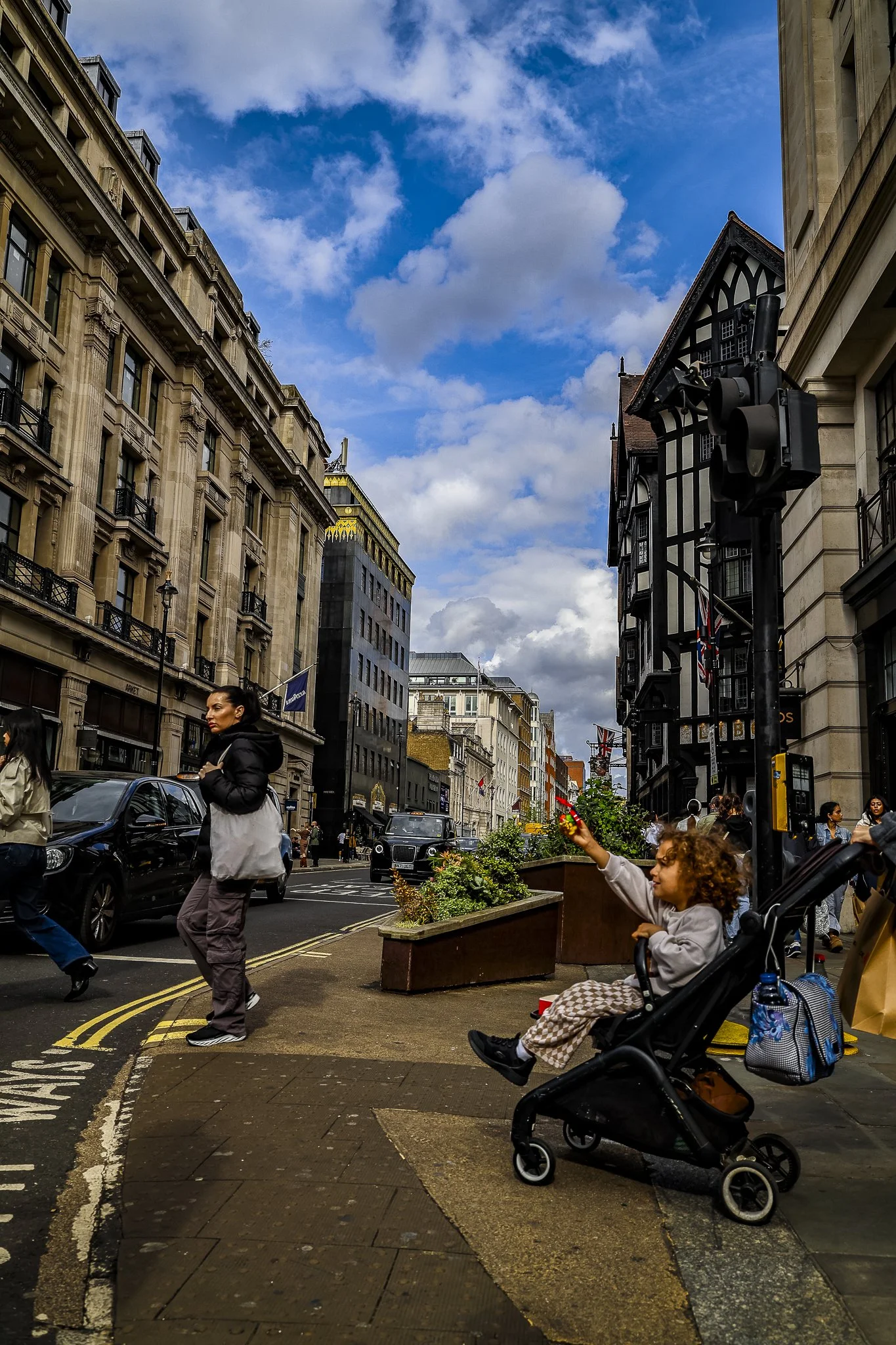 City street scene with historic and modern buildings, people walking and standing, a child sitting in a stroller reaching out, and cars parked along the street under a blue sky with scattered clouds.