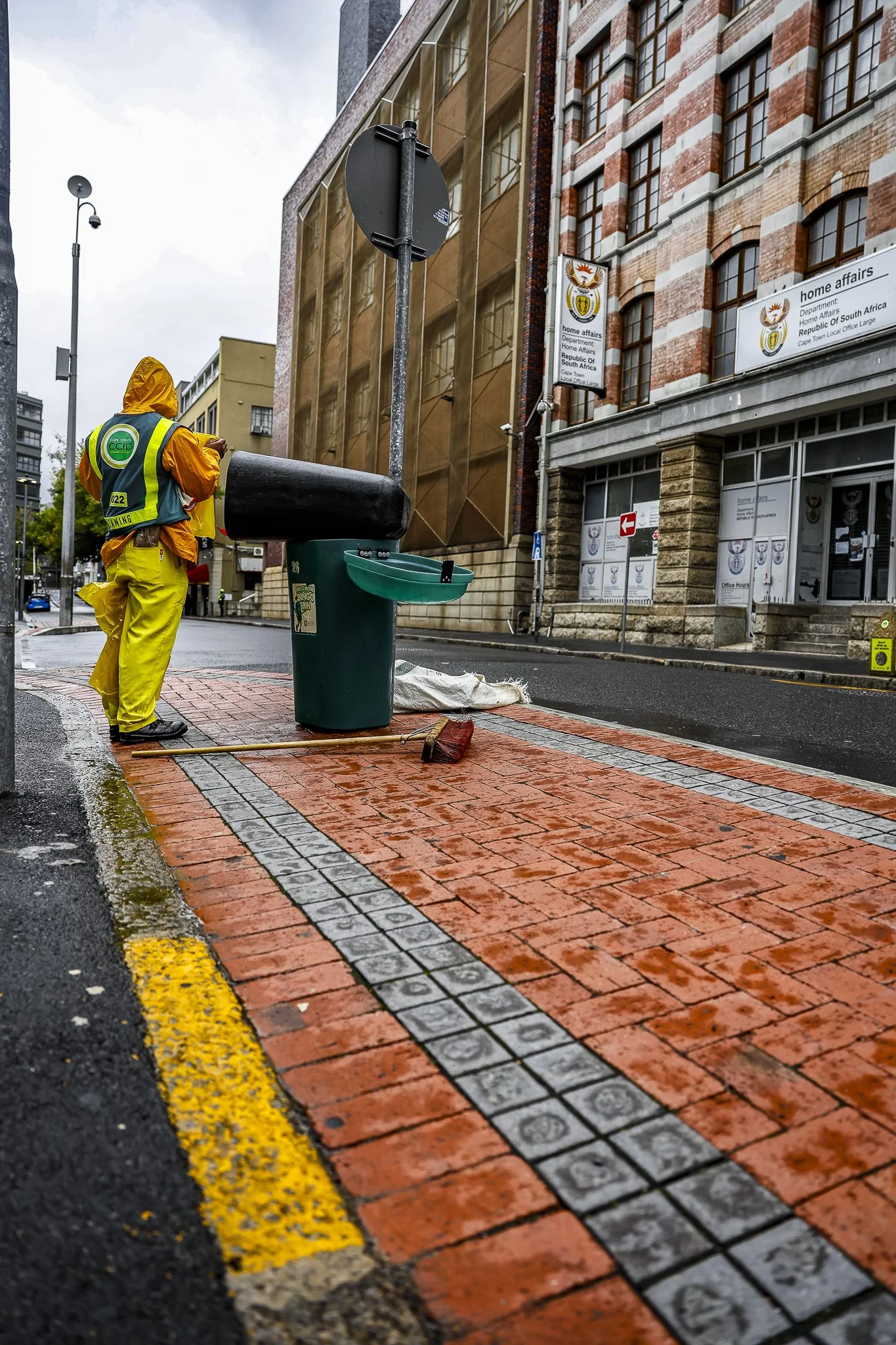 A city street corner with a worker in yellow rain gear, a green garbage bin, and a brick-paved sidewalk with a yellow line marking the edge. There are buildings with signs, including one for home affairs, and lampposts, under cloudy weather.