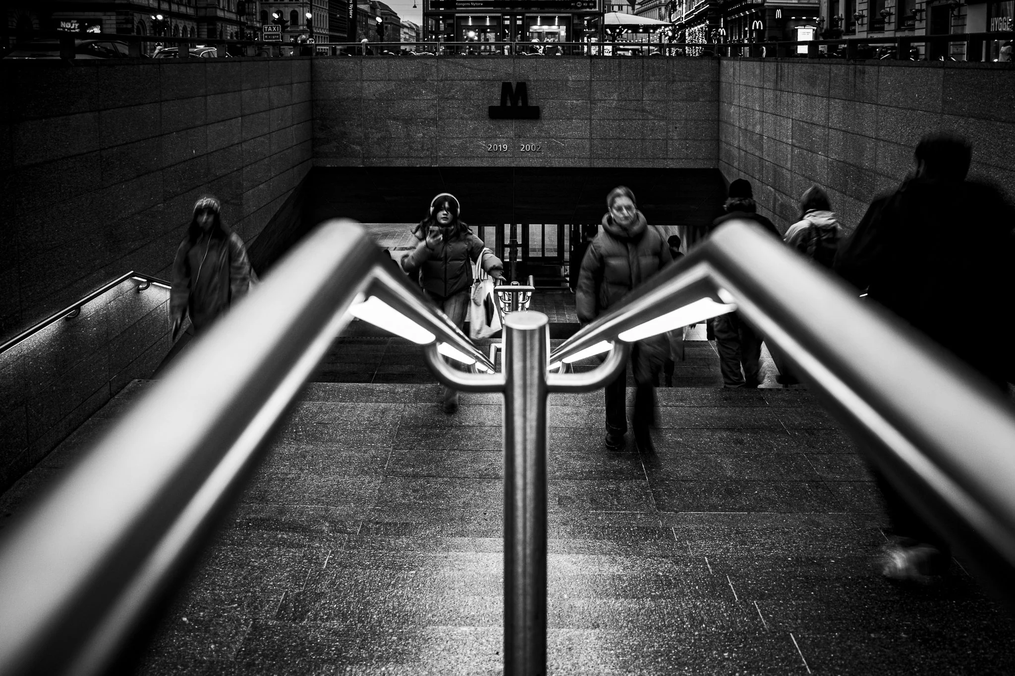 Black and white photo of people descending a staircase in an urban area, with a subway entrance at the bottom, visible signs, cars, and city buildings in the background.