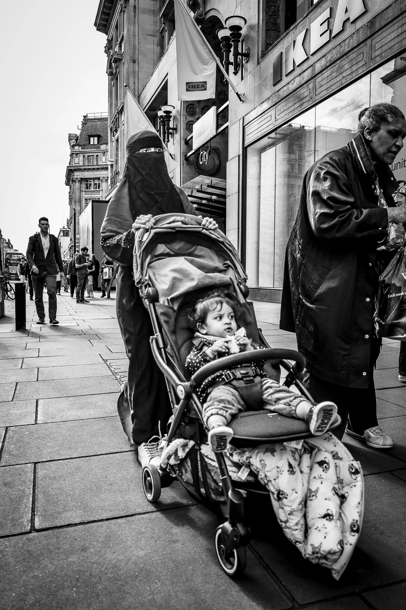 Black and white street scene with people walking on the sidewalk in front of a store with IKEA signage. A woman wearing a niqab pushes a stroller with a young child in pajamas, and other pedestrians are visible in the background.